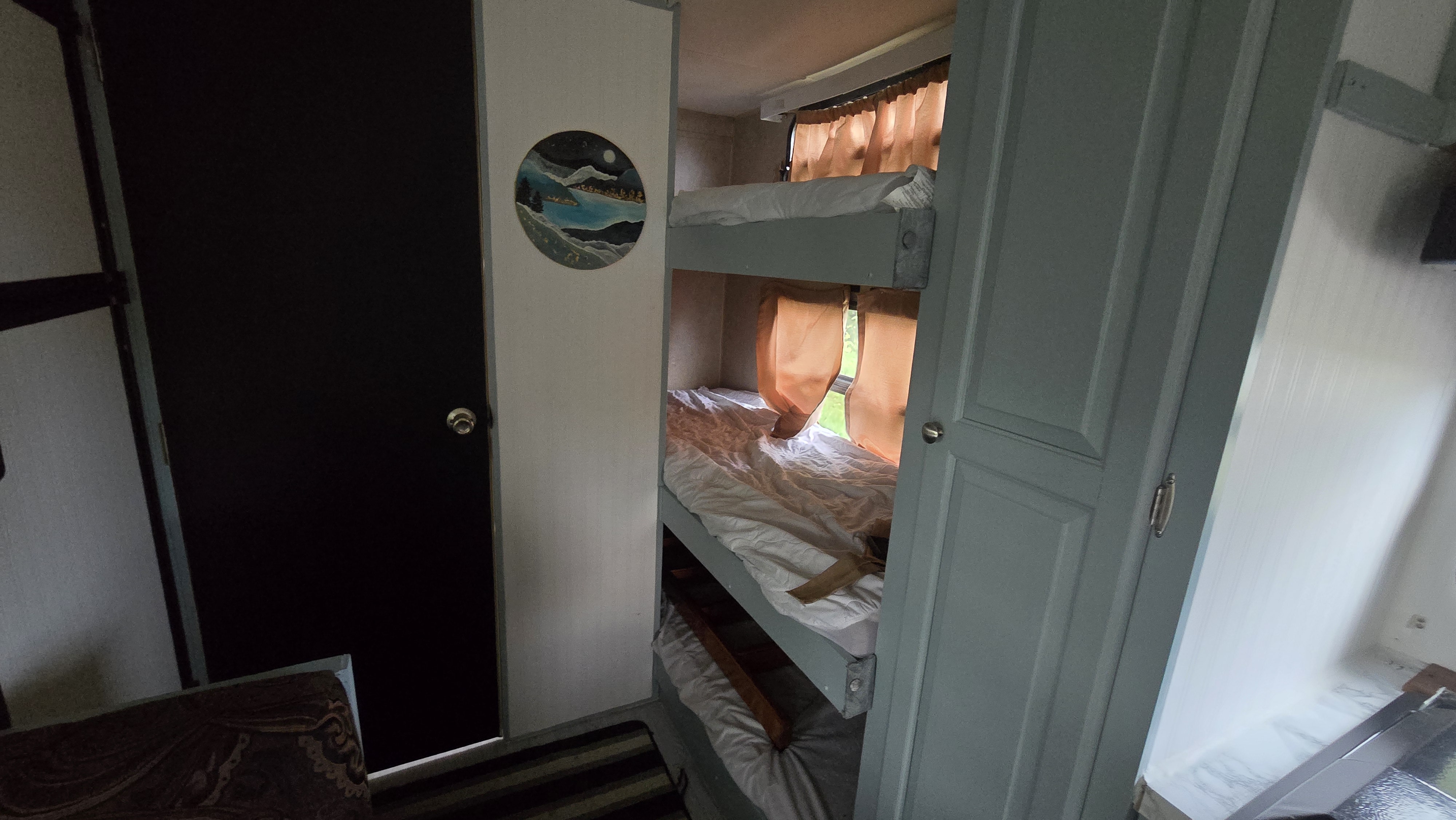 Inside an RV rental: two bunk beds with white sheets, orange window curtains, a dark door, and a round landscape painting.. Forest River Rockwood Mini Lite 2012