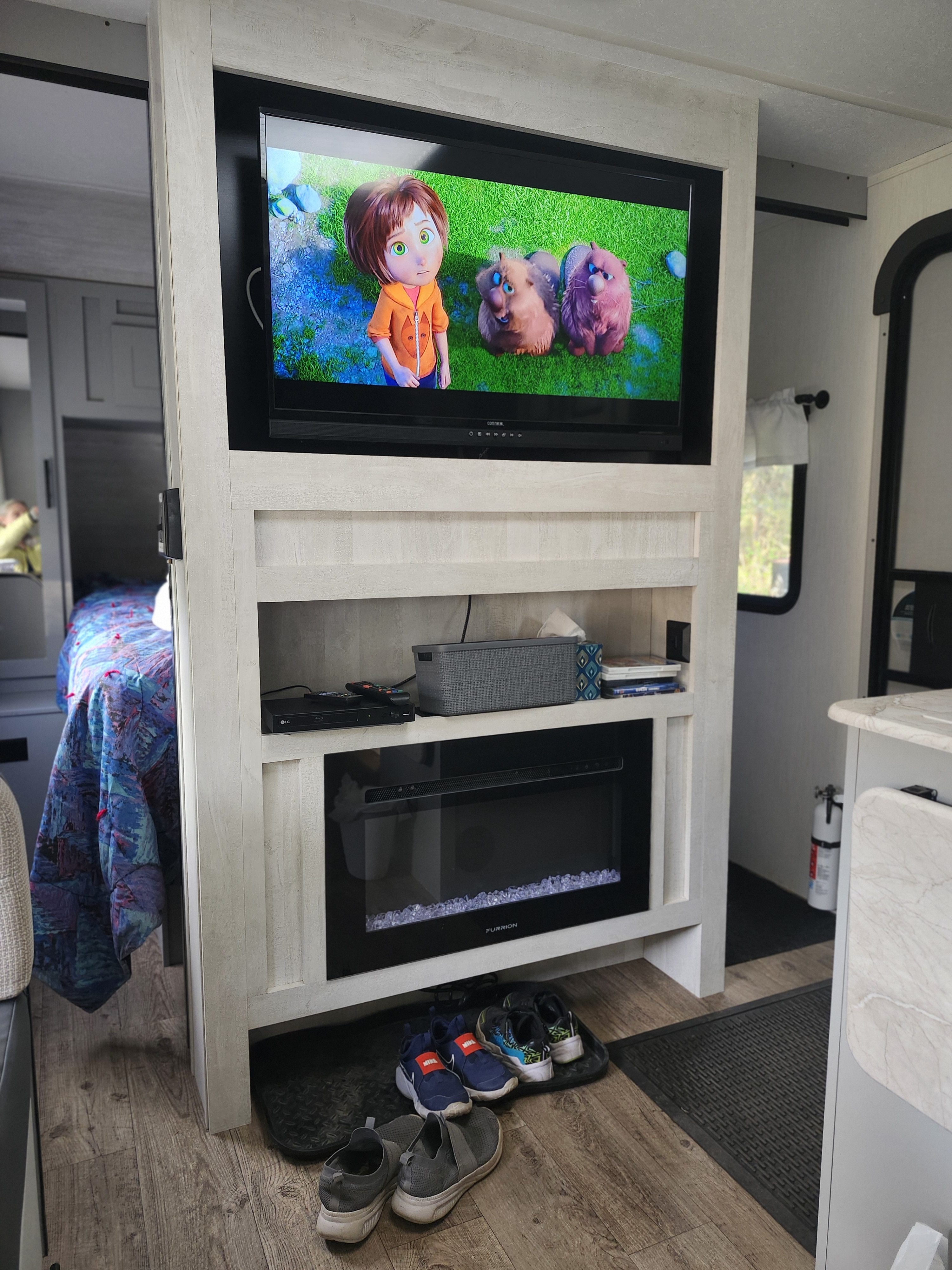 This RV rental's living area features a TV displaying cartoons, an electric fireplace, shelves, and shoes neatly placed on mats.. Forest River Vibe 2023