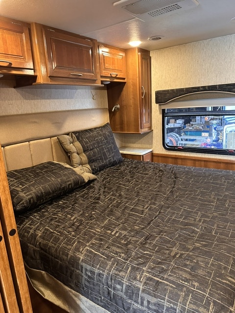 Cozy RV rental bedroom featuring dark patterned bedding, ample wood cabinetry, and a window view, ideal for travel.. Forest River Coachmen Mirada 2018