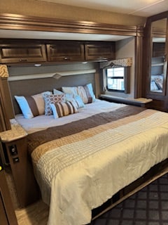 Spacious RV rental bedroom featuring a large bed with striped bedding, dark wood overhead cabinets, and a side window.. Thor Hurricane 2020
