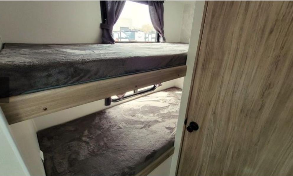 Cozy RV rental bunk beds. Upper bunk has a window with purple curtains. Lower bunk visible, both with soft gray bedding.. Winnebago Access 2025