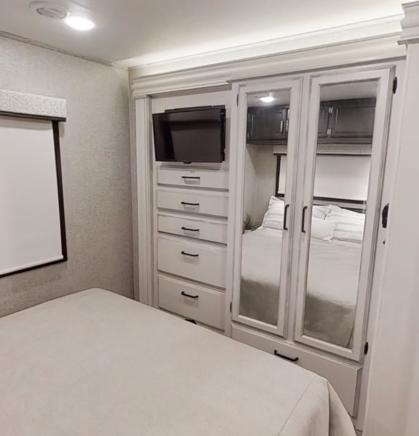 This RV rental bedroom features a bed, a window with shade, and a white built-in unit with a TV, drawers, and mirrored wardrobe doors reflecting the bed.. Jayco Alante 2023