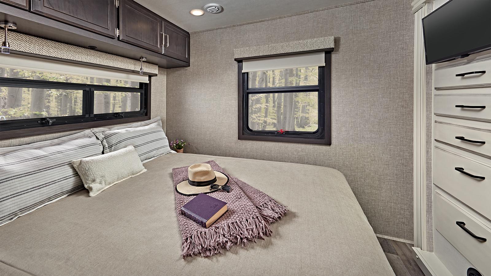 This cozy RV rental bedroom features a comfy bed, scenic window views of a forest, overhead cabinets, and a dresser.. Jayco Alante 2023