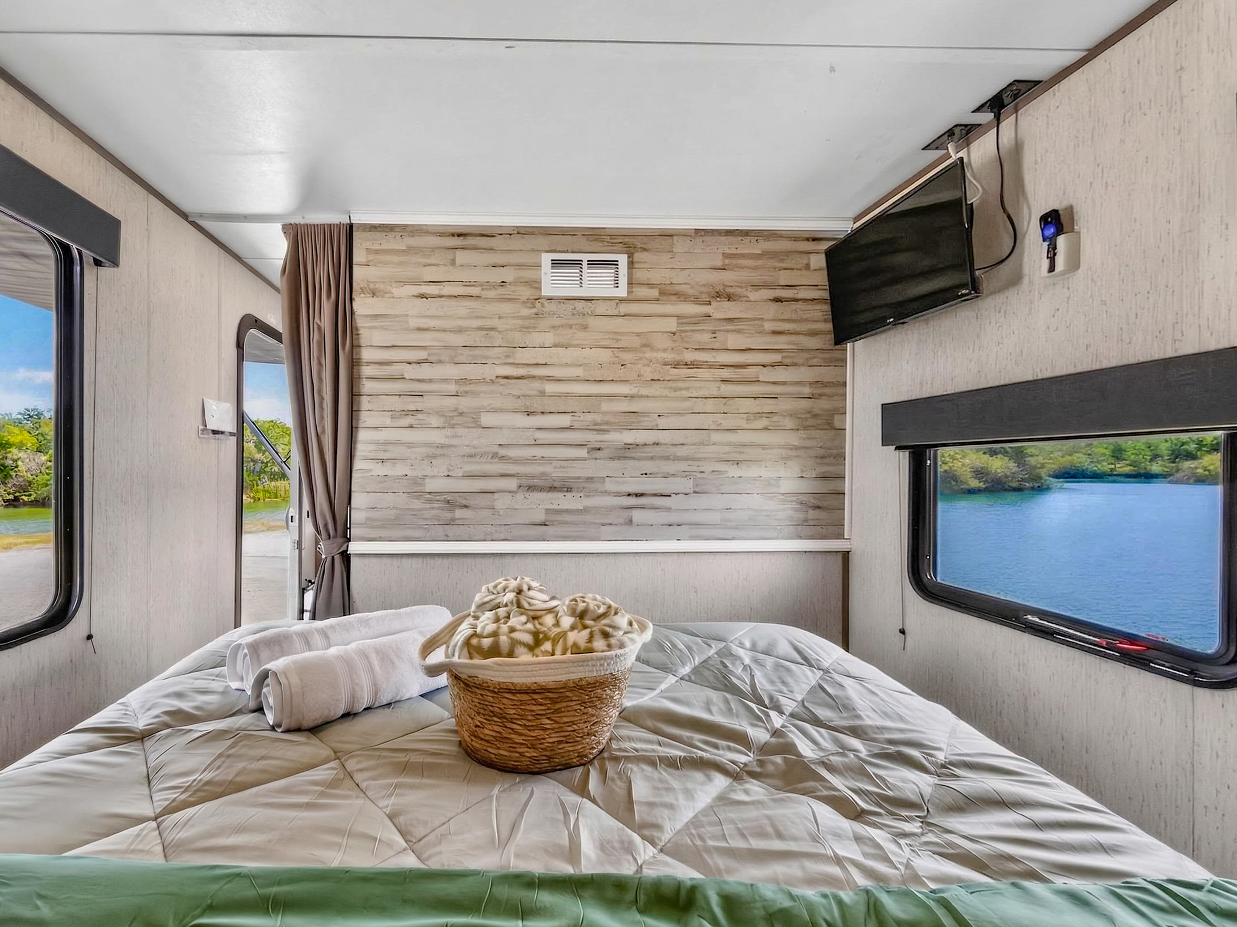 RV rental bedroom with a comfortable bed, towels, and basket. Enjoy lake views from large windows, a wood accent wall, and a TV.. Dutchmen Coleman Lantern LT 2022