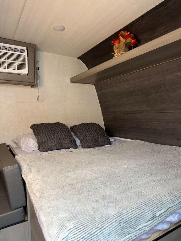 RV rental interior showing a cozy bedroom: bed with textured blanket/pillows, wall AC unit, and shelf with decor.. Keystone RV Hideout Sport 2024