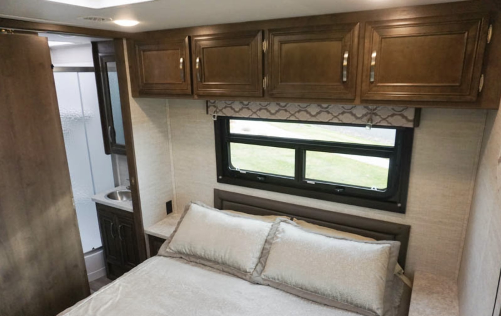 Compact RV rental bedroom with a comfy bed, window, overhead storage, and partial view of the attached bathroom's sink.. Jayco Redhawk 2020