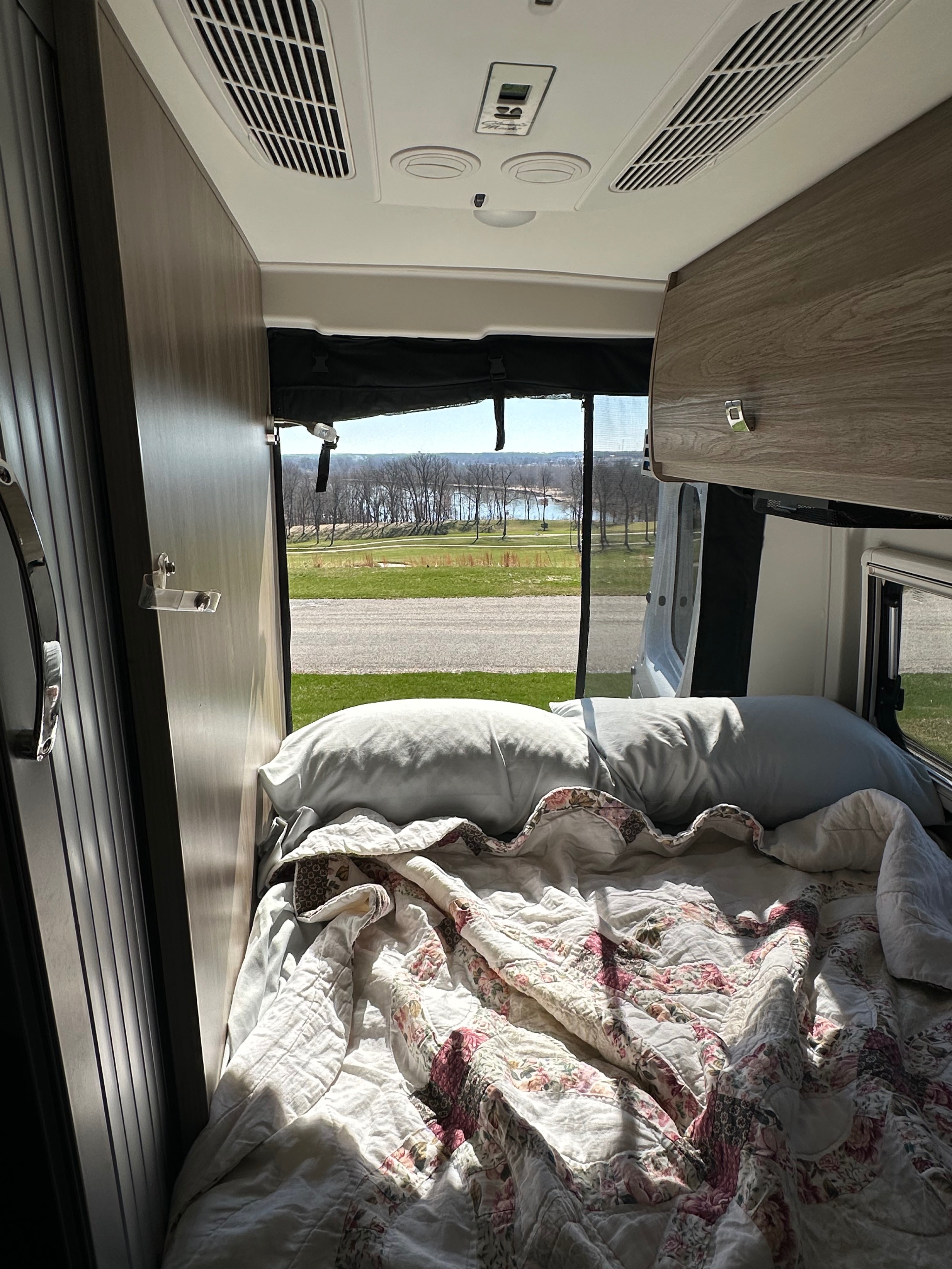 Inside an RV rental: a sunlit bed with floral quilt, offering a scenic view of a lake through bare trees. Perfect for adventure.. Winnebago Travato 2023