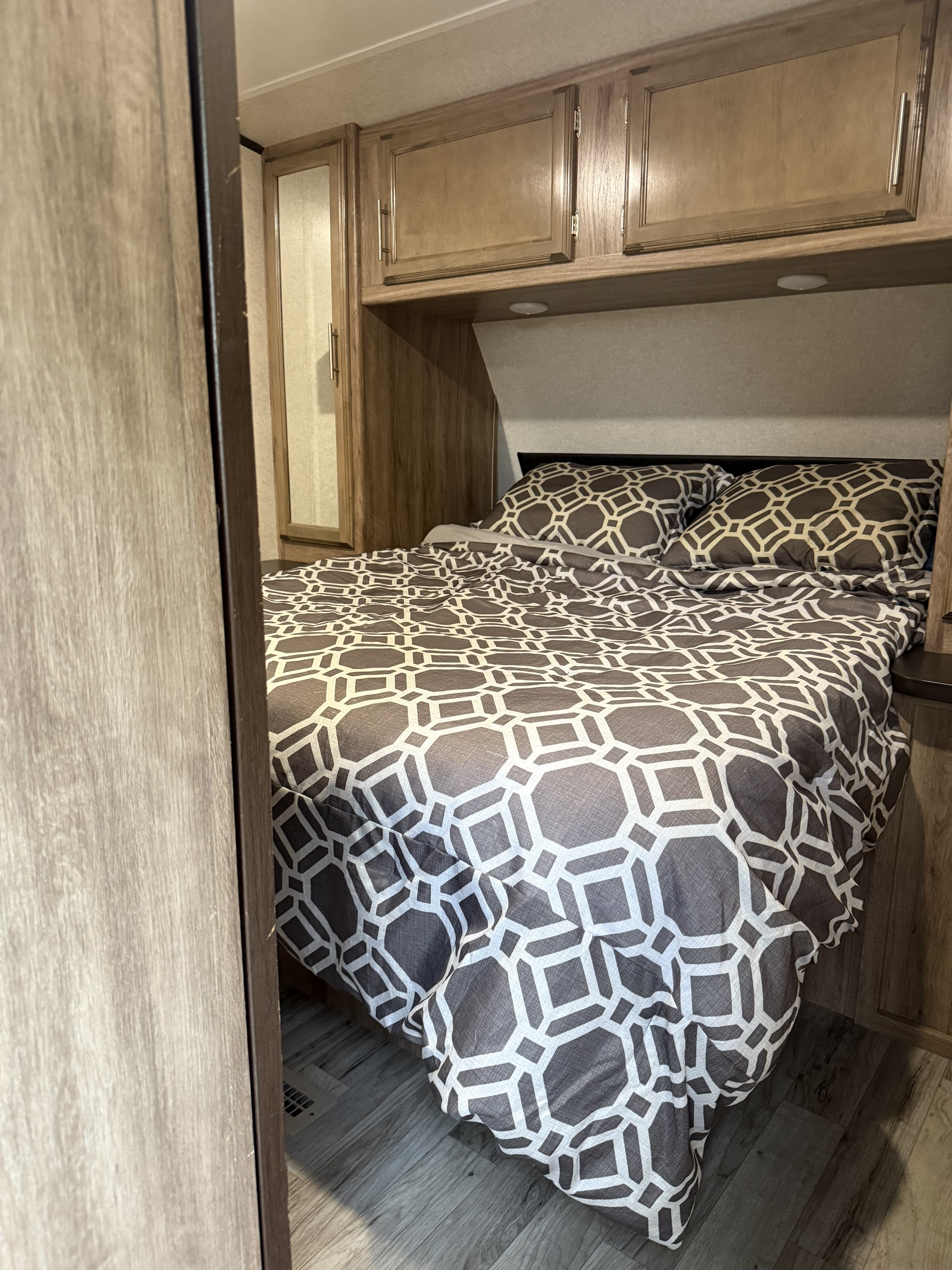 Cozy RV rental bedroom with geometric patterned bedding, overhead wooden cabinets, and wood-look flooring for a compact space.. Jayco Jay Feather 2019