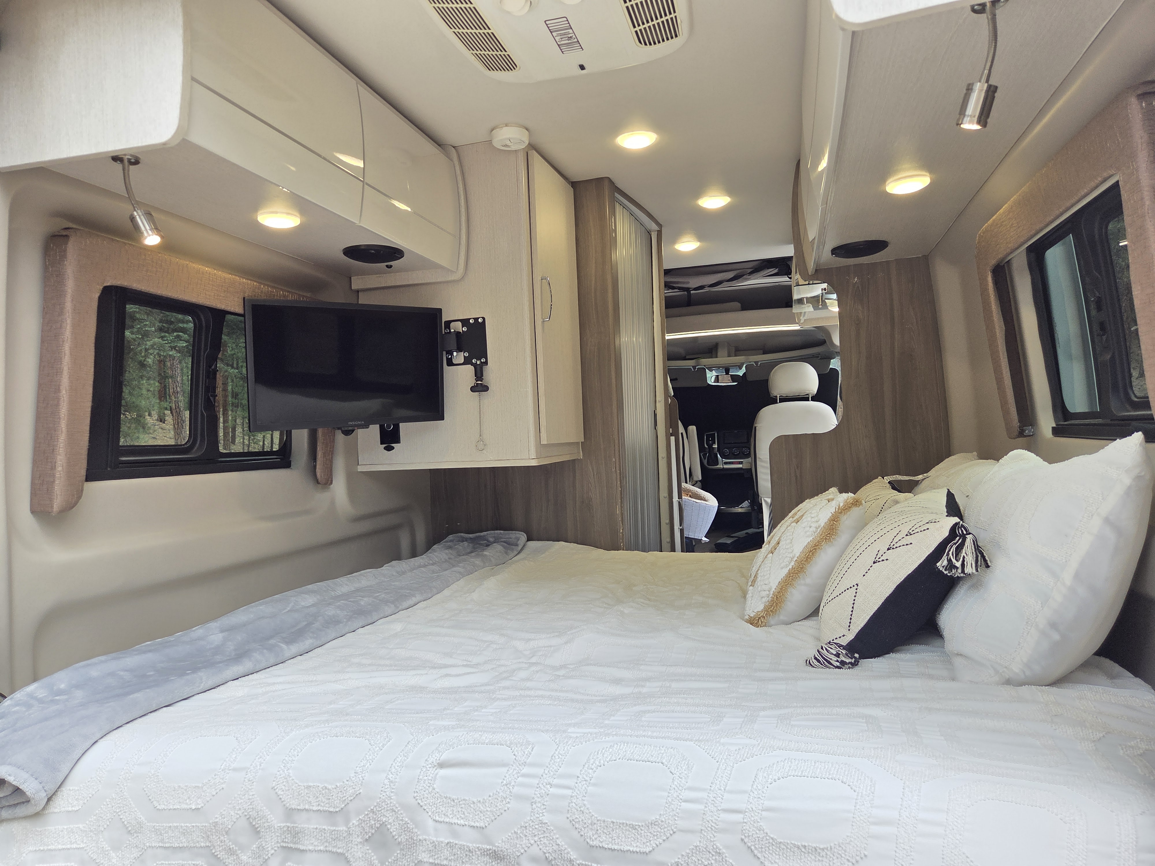This RV rental offers a comfortable bedroom with a TV, plush pillows, and peaceful forest views from the window.. Thor Tellaro 2022