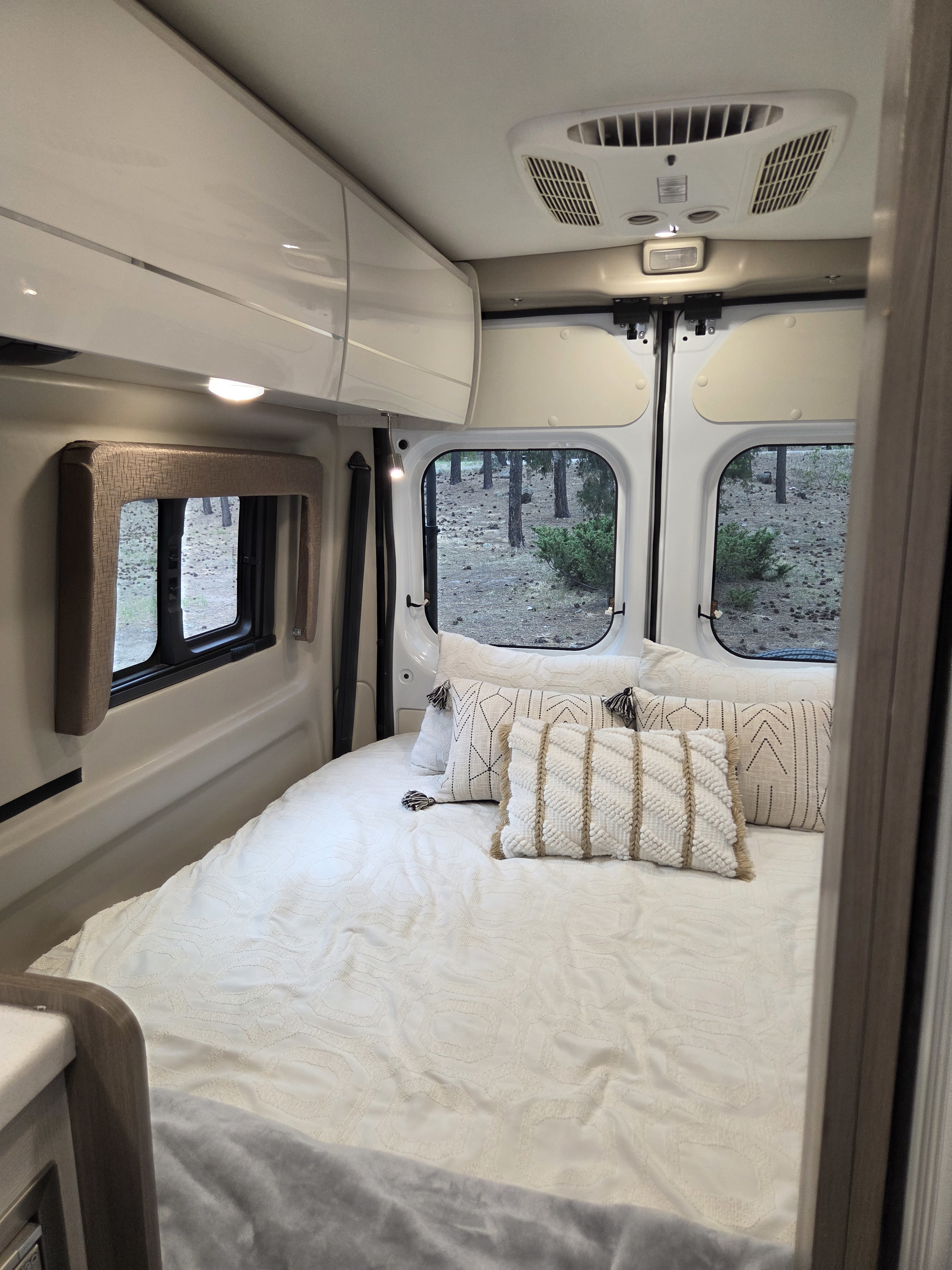 Cozy rv rental bedroom interior with plush white bedding, decorative pillows, and scenic window views of the outdoors.. Thor Tellaro 2022