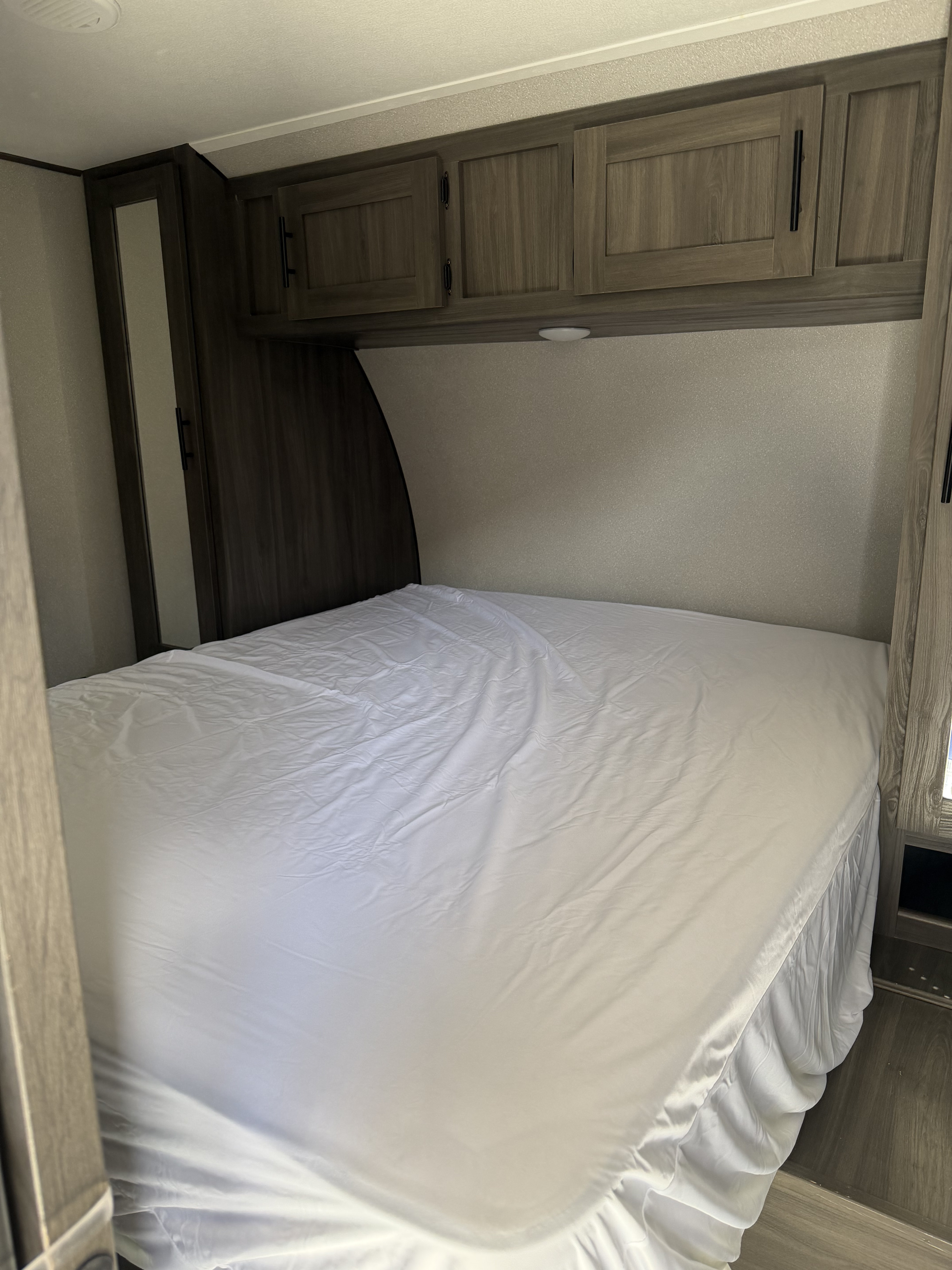 Cozy RV rental bedroom featuring a bed with white sheets, overhead wooden cabinets, and a narrow wardrobe, ready for travel.. Forest River Shasta 2021