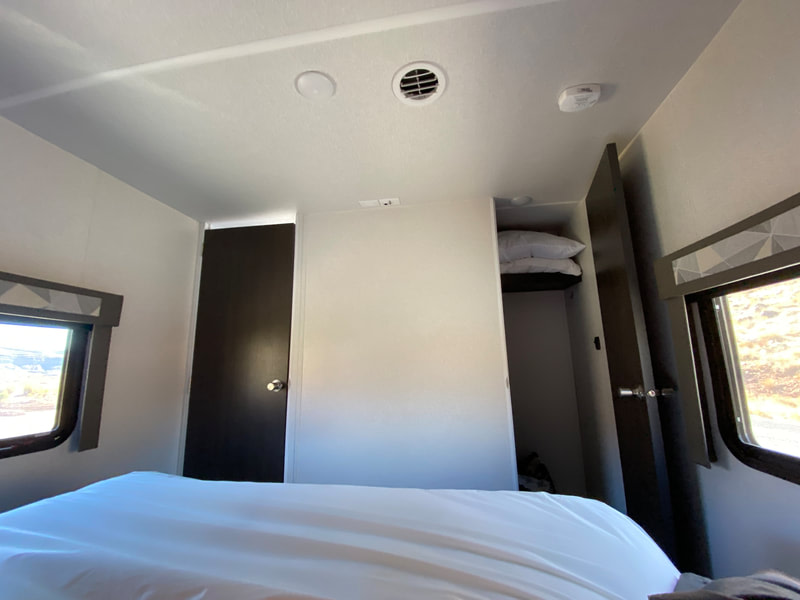 Cozy RV rental bedroom features a white bed, open closet with pillows, and windows revealing outdoor views.. Forest River Salem 2020