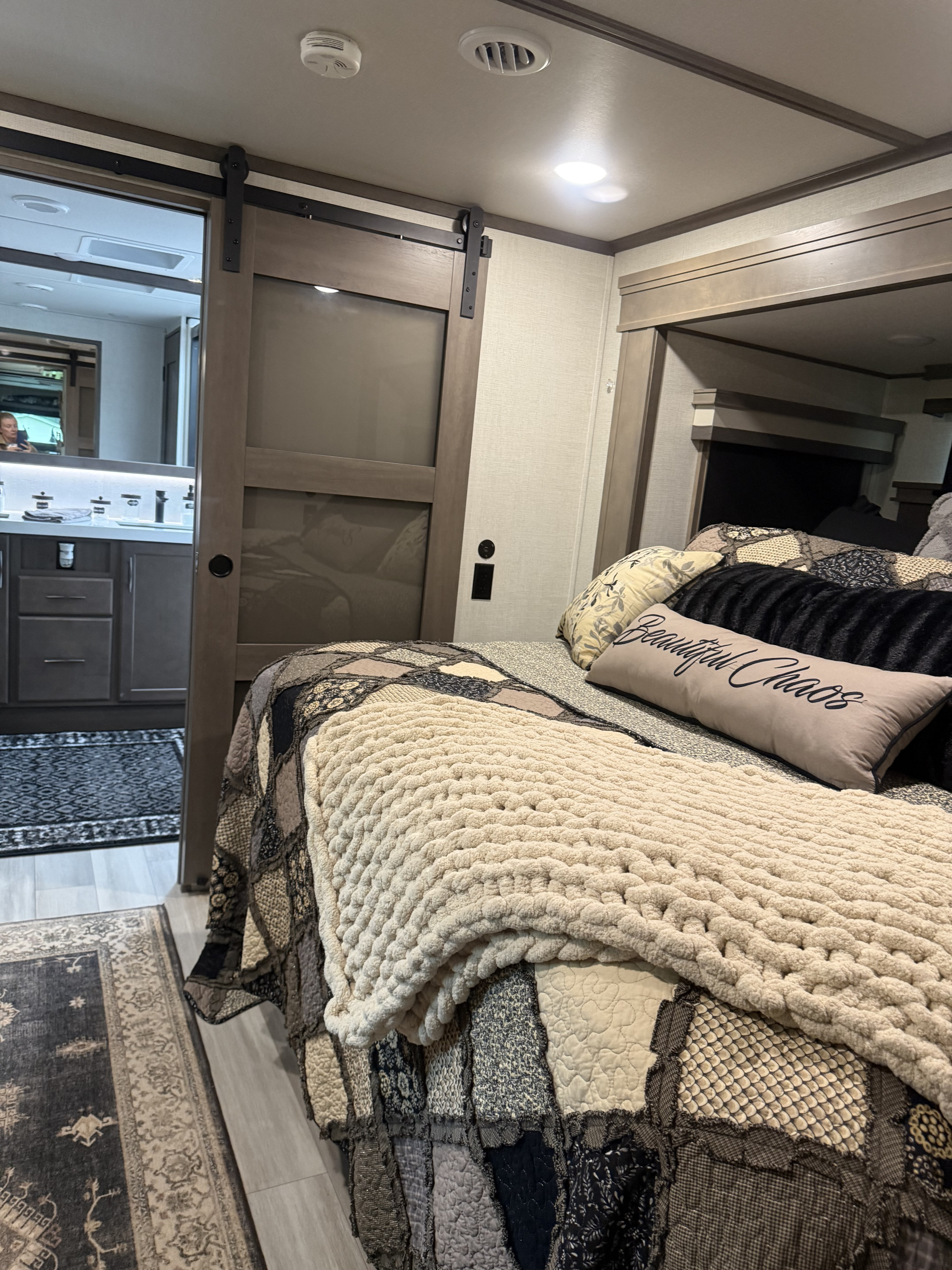 Inside a spacious RV rental: a cozy bedroom with a patchwork quilt, plush throw, and barn door entry to the bathroom.. Grand Design Solitude 2023