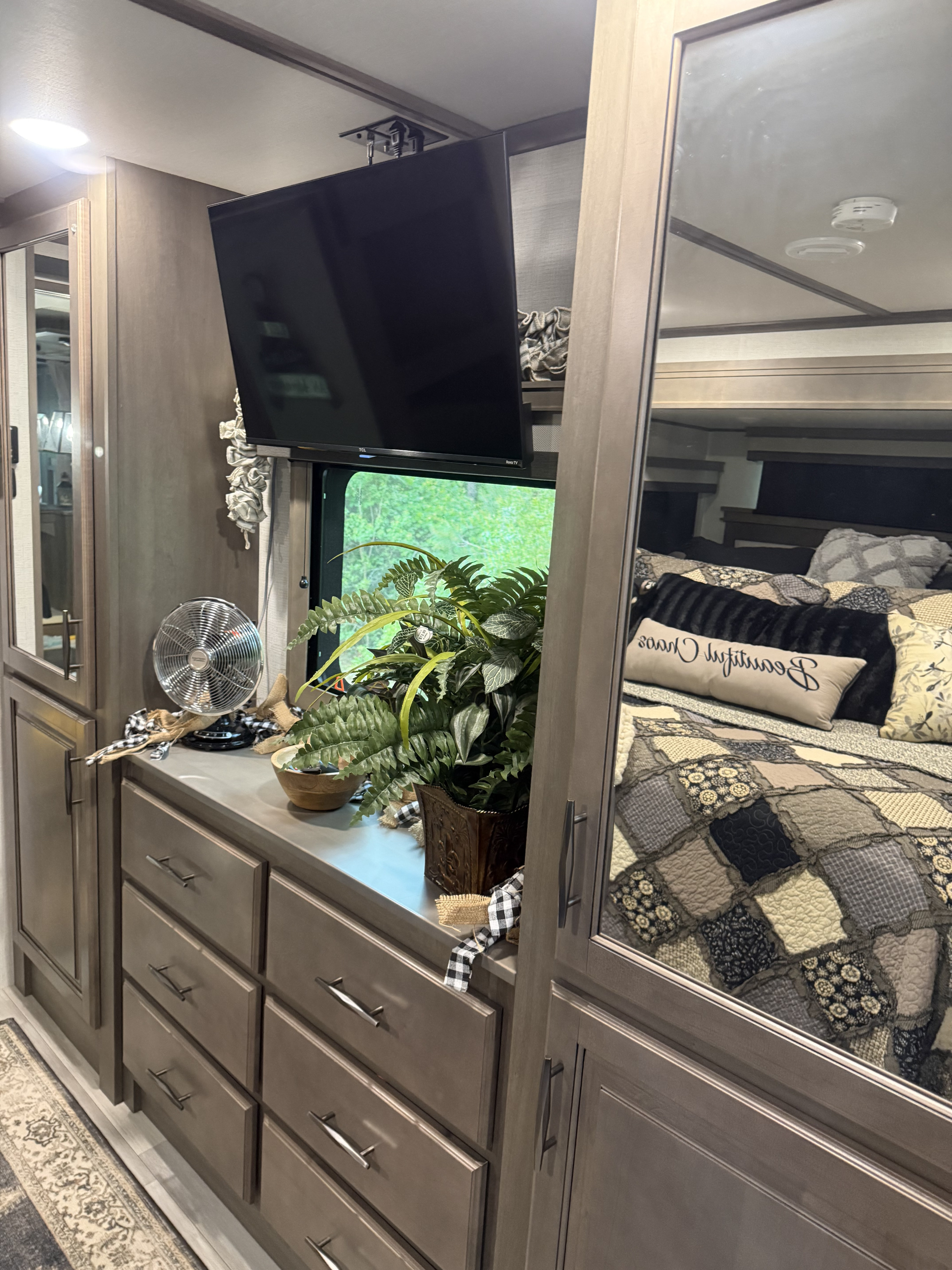 Inside a cozy RV rental, a bedroom area with a TV, ample storage drawers, and decorative touches. A mirrored cabinet reflects the comfortable bed.. Grand Design Solitude 2023