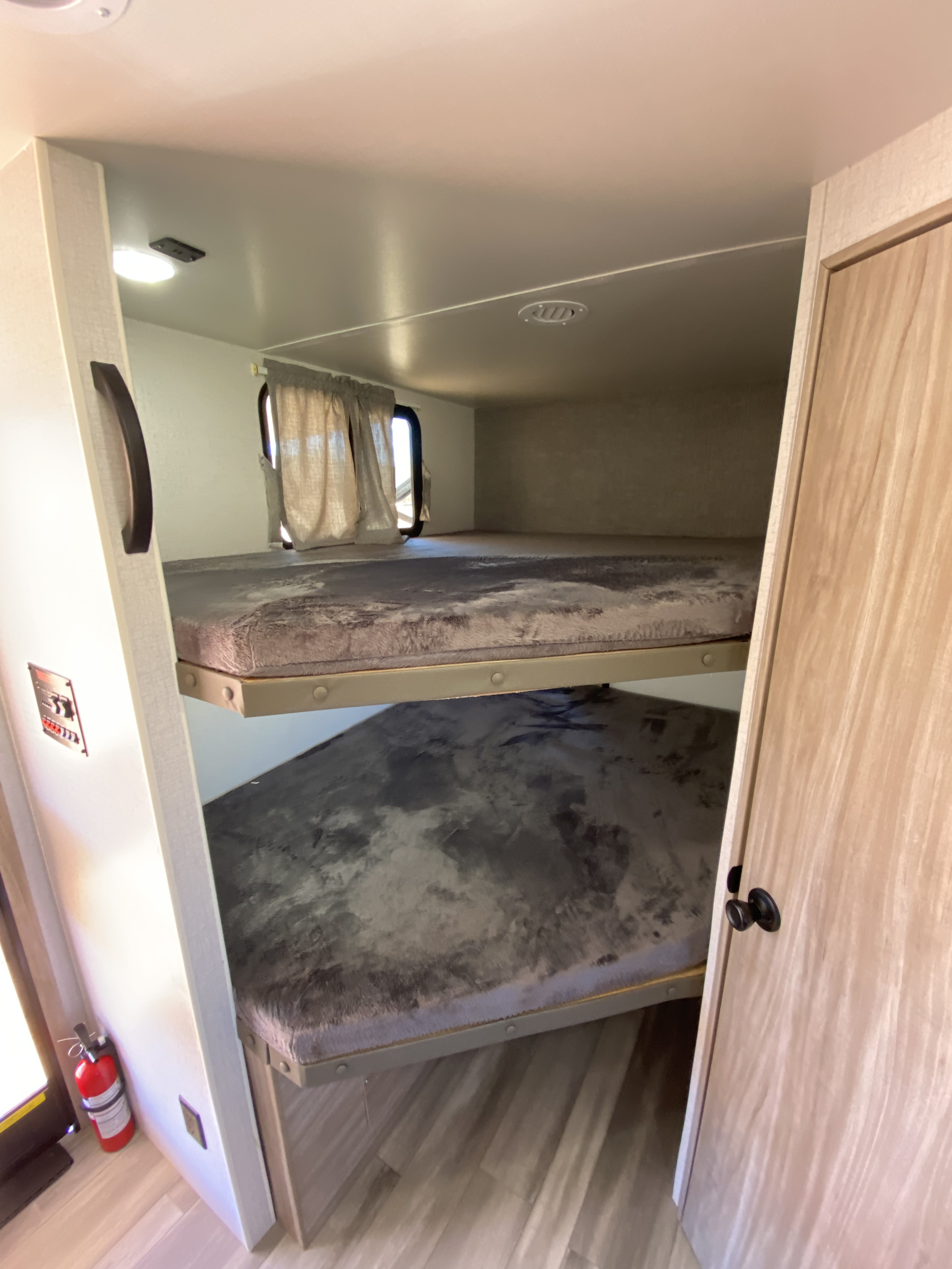 An RV rental interior featuring stacked bunk beds with plush grey mattresses, a window with curtains, a control panel, and a fire extinguisher.. Winnebago Minnie Winnie 2022