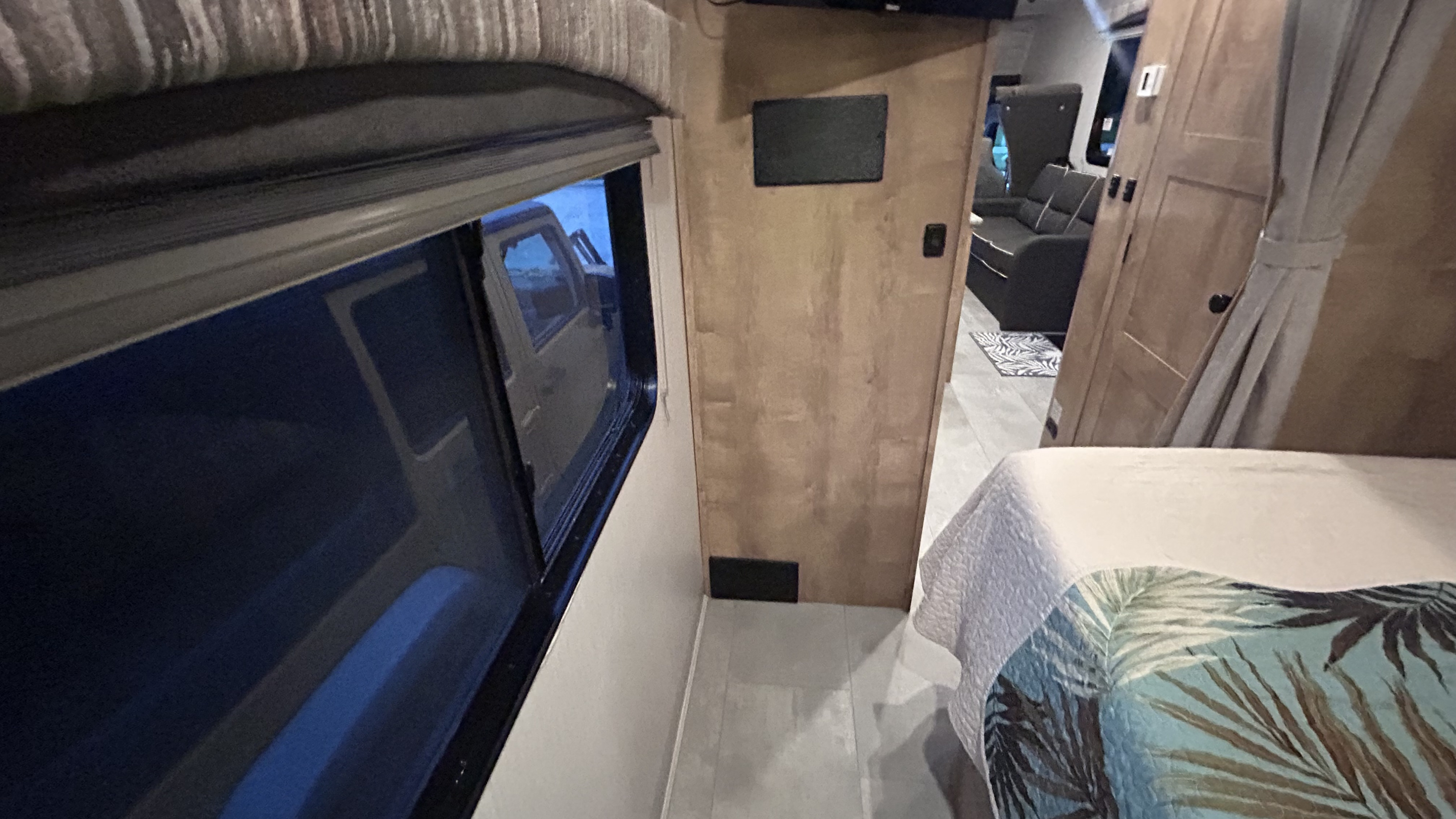 RV rental interior shows a bedroom with a tropical bedspread, a window reflecting a vehicle, and a living area beyond.. Forest River Coachmen Leprechaun 2022