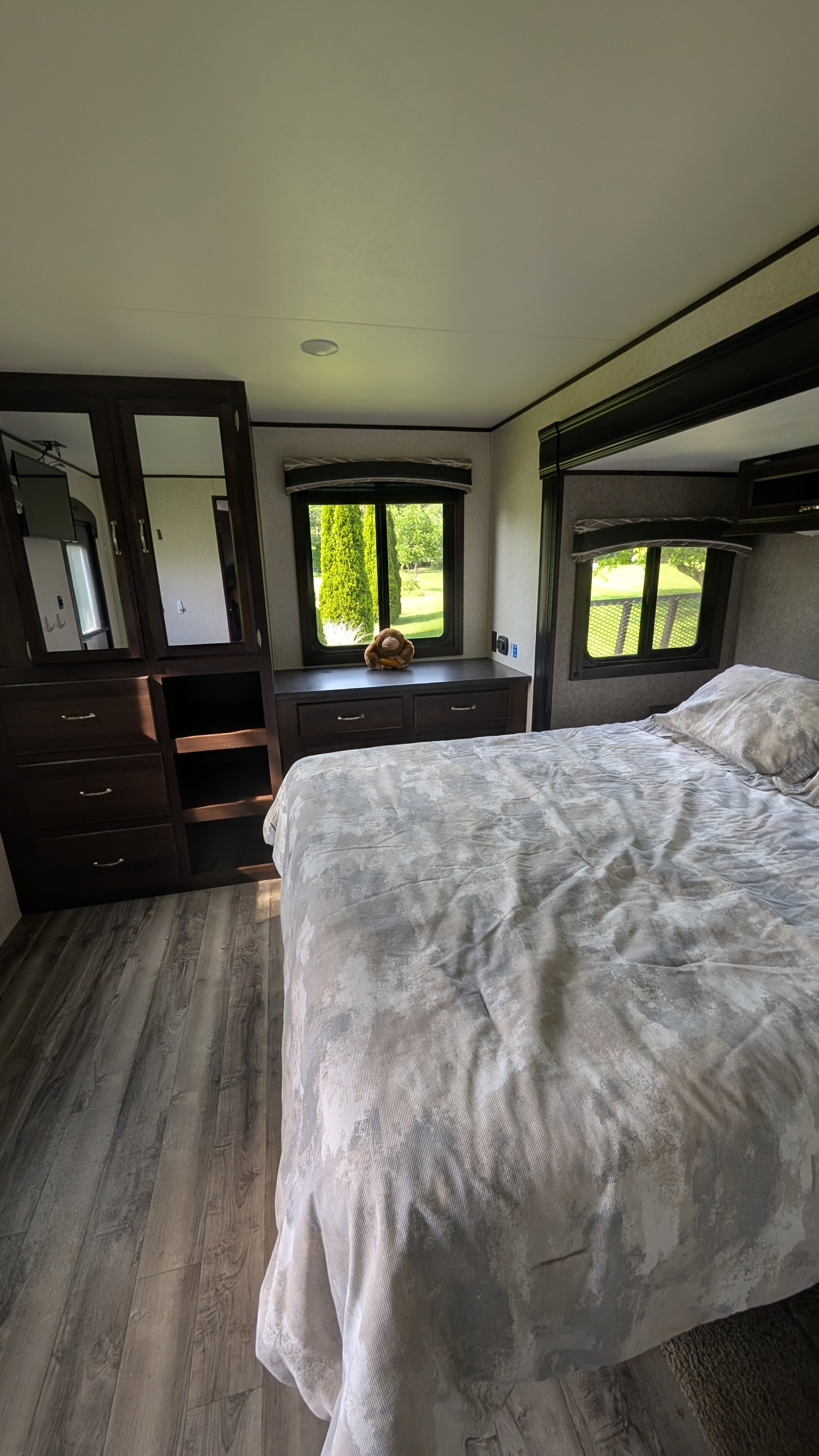 Interior of an RV rental bedroom featuring a patterned bed, dark wood furniture, and bright windows with green views.. Jayco Jay Flight 2017