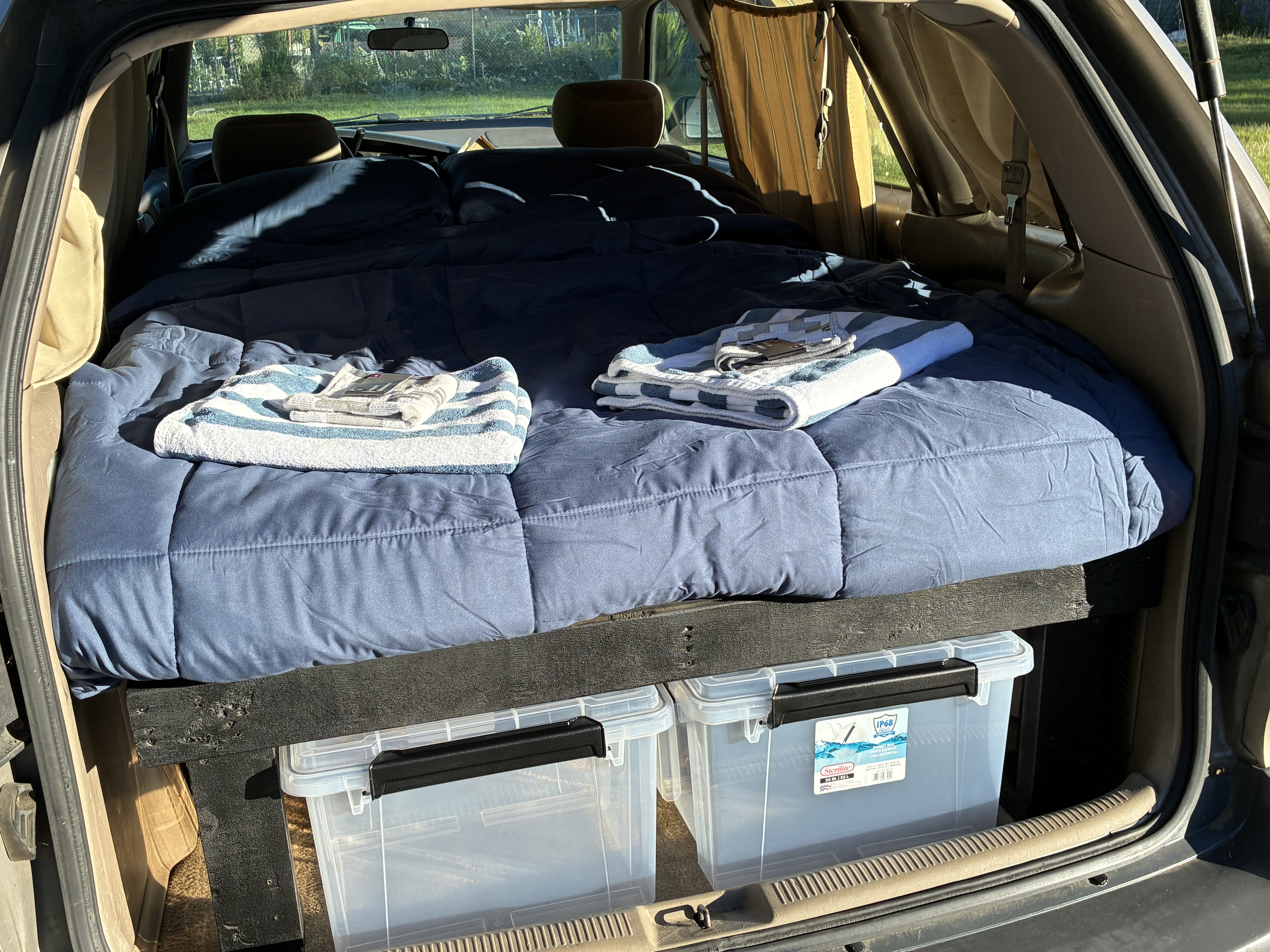 Inside a car camping setup: a comfy bed on a wooden platform over storage bins, a budget-friendly RV rental alternative.. Toyota Matilda 2000