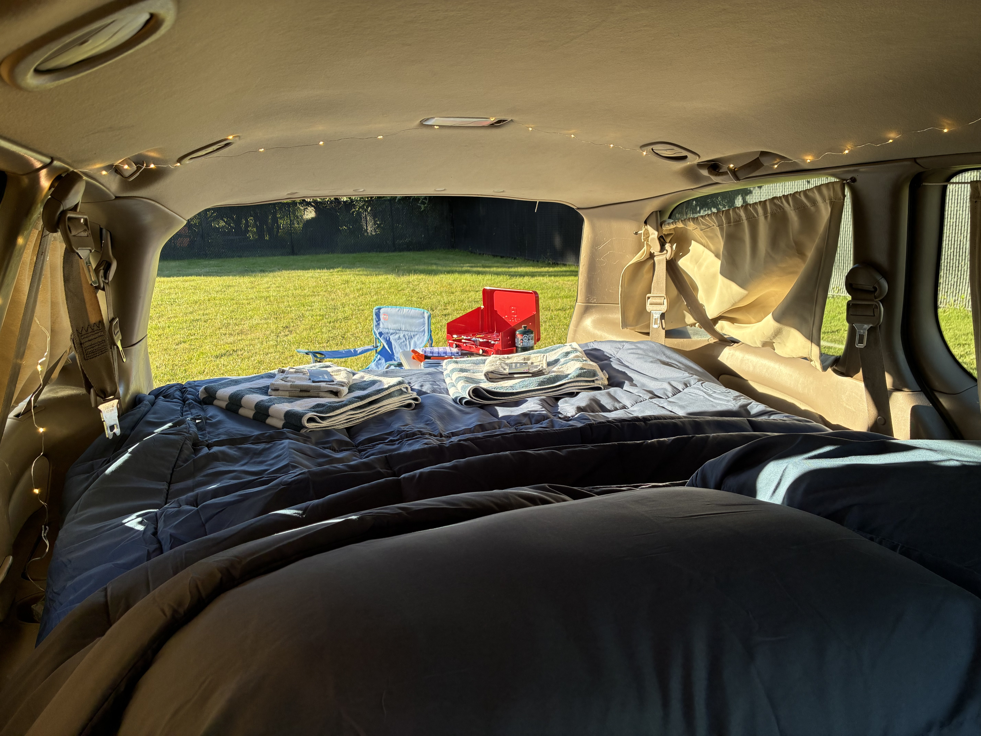 Cozy van camping setup inside an RV rental with a comfortable bed, string lights, and an outdoor view of a grassy yard with a stove.. Toyota Matilda 2000
