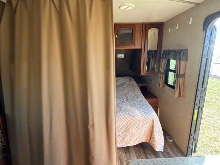 Cozy RV rental bedroom with a drawn curtain, comfortable bed, cabinets, and open door showing nature. Perfect for travel.. Heartland RVs North Trail 2014