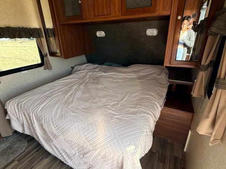 Cozy RV rental bedroom: features a large, comfortable bed, window with a view, and mirrored cabinet reflecting a person.. Heartland RVs North Trail 2014