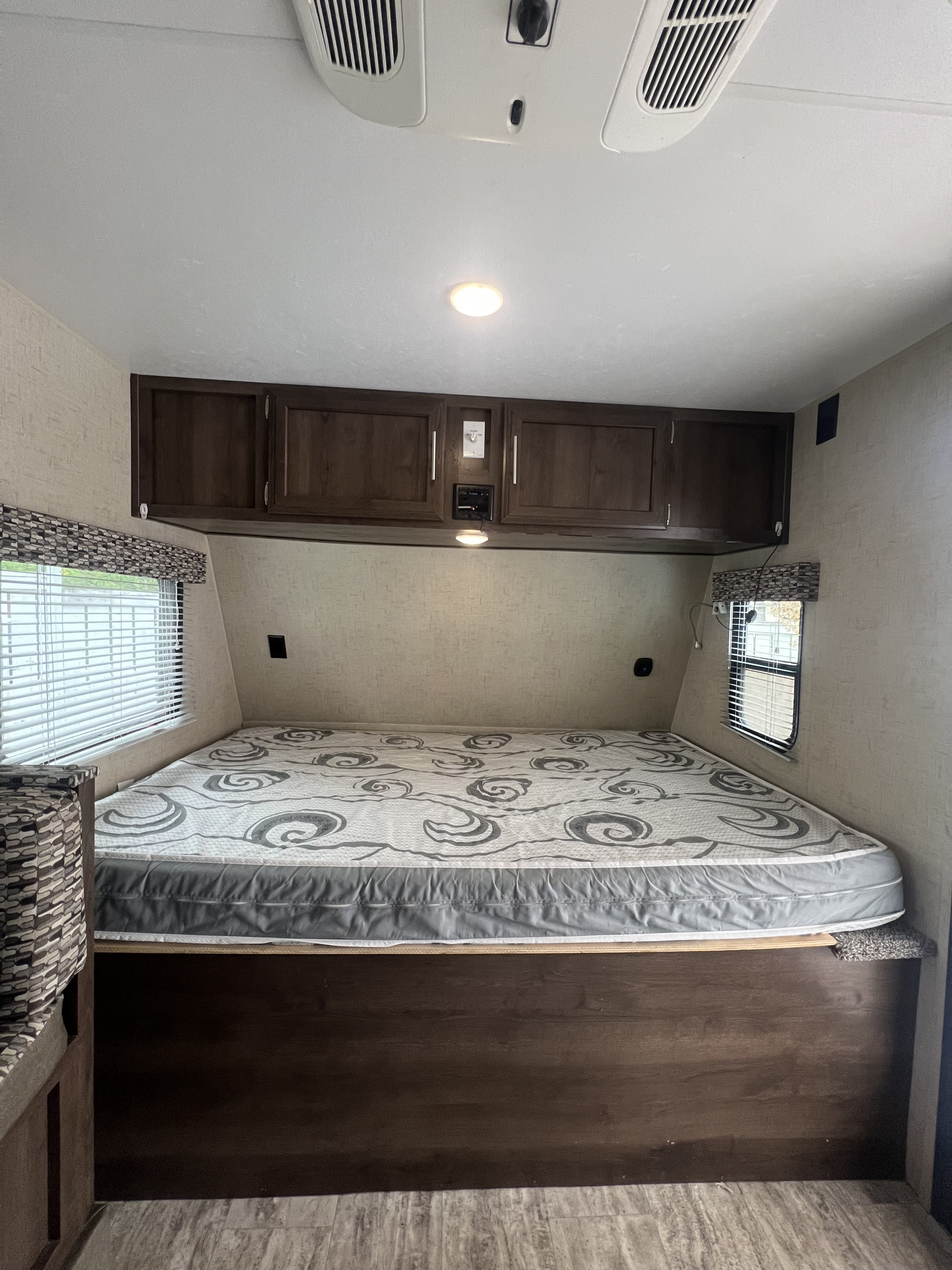 RV rental bedroom featuring a patterned bed, dark wooden overhead cabinets, and windows with blinds for a comfortable stay.. KZ Sportsmen Classic 181BH 2018