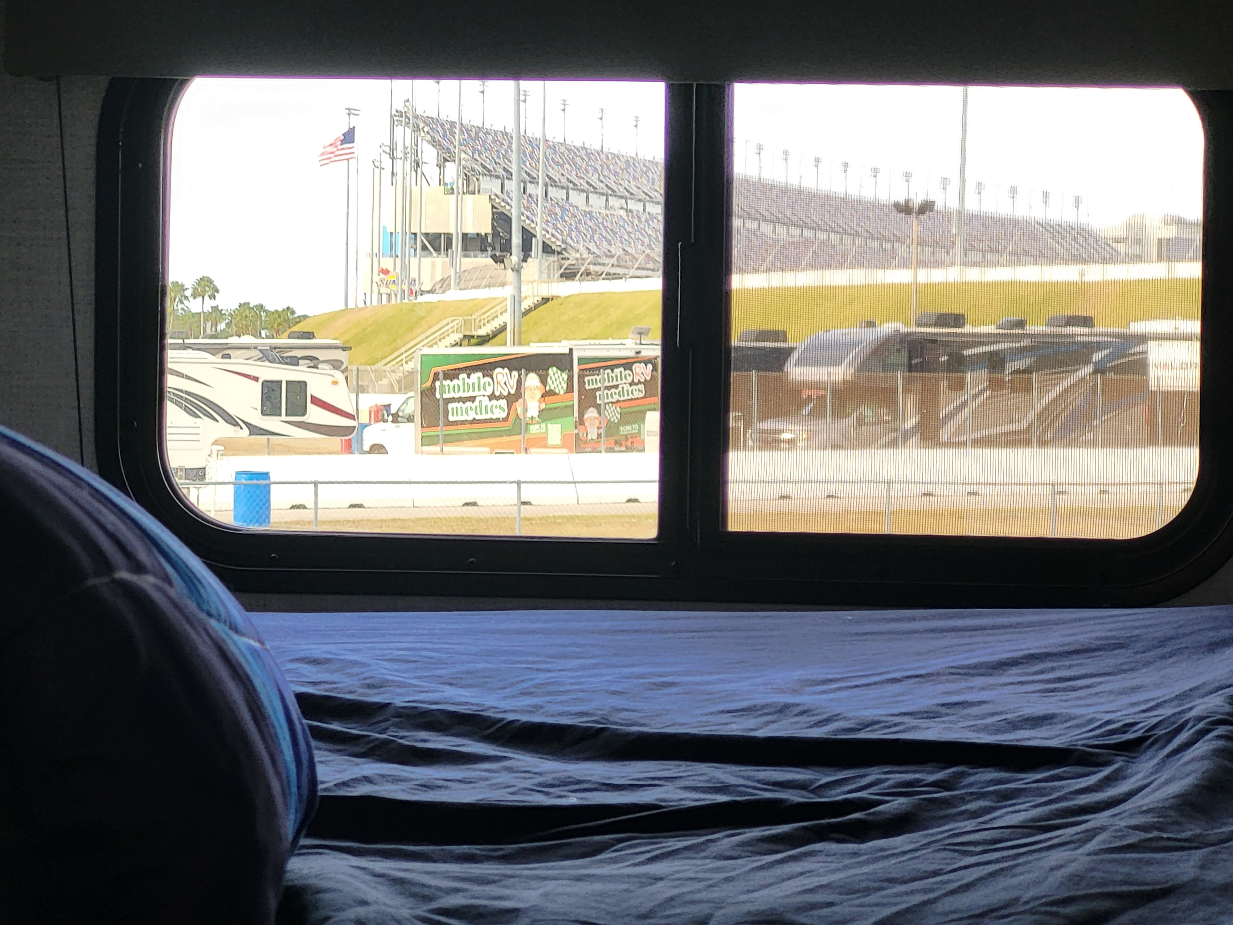 View from an RV rental window looking out at racetrack grandstands, parked RVs, and "Mobile RV Medics" vehicles.. Grand Design Transcend Xplor 24BHX 2025