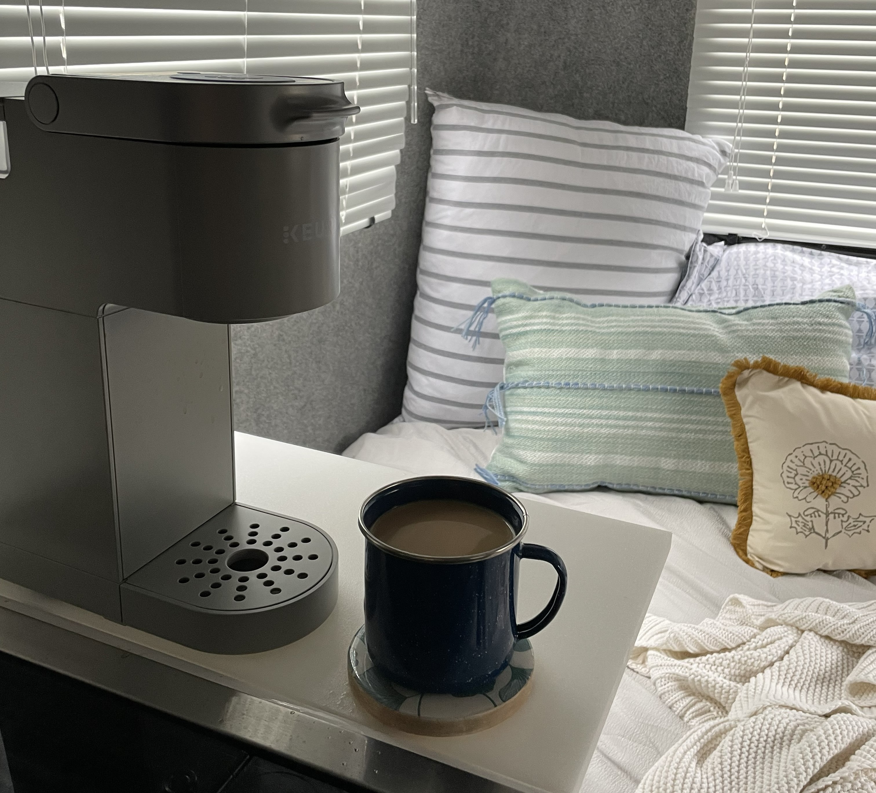 RV rental comfort: A gray Keurig, dark coffee mug, and various pillows create a cozy scene next to window blinds.. Casita Freedom 2024