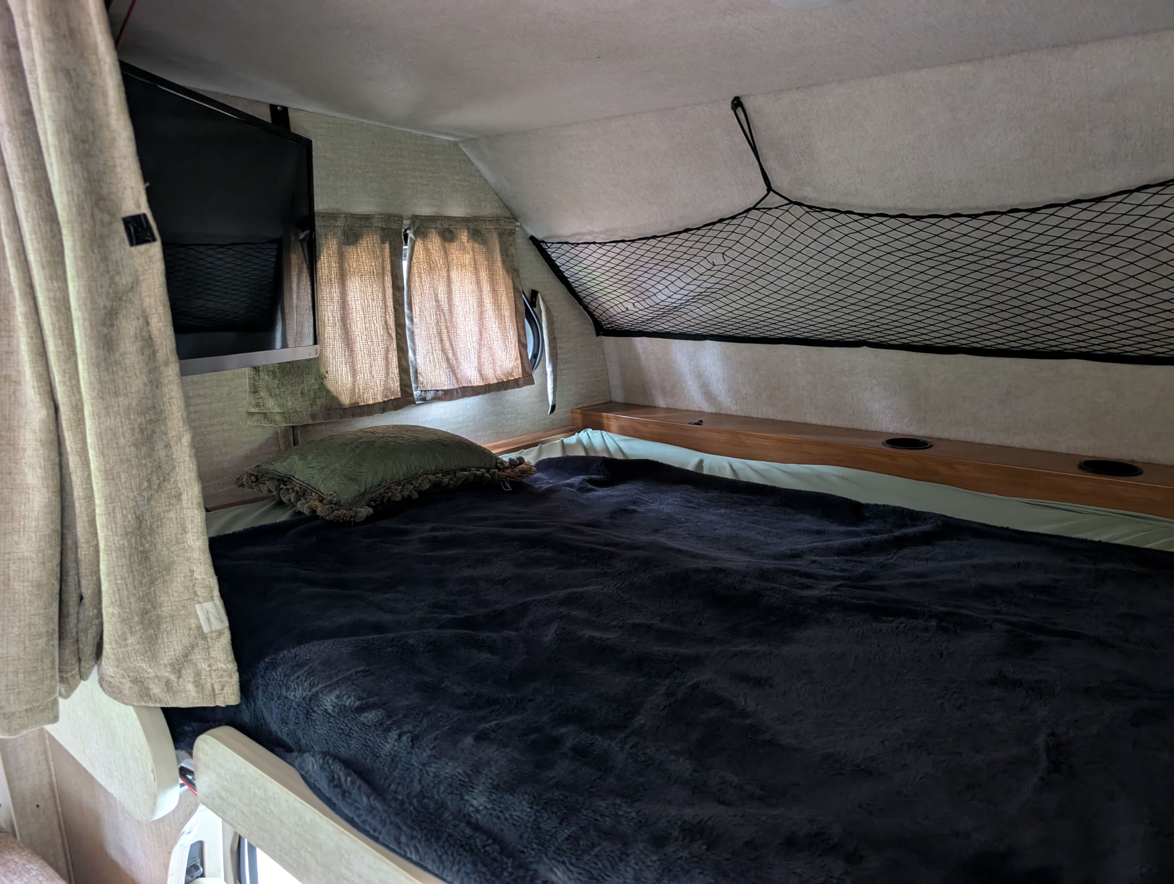 Inside this RV rental: a cozy bed with a dark blanket, TV, storage net, and windows with curtains for a comfortable trip.. Tioga Tioga Motorhome 2008
