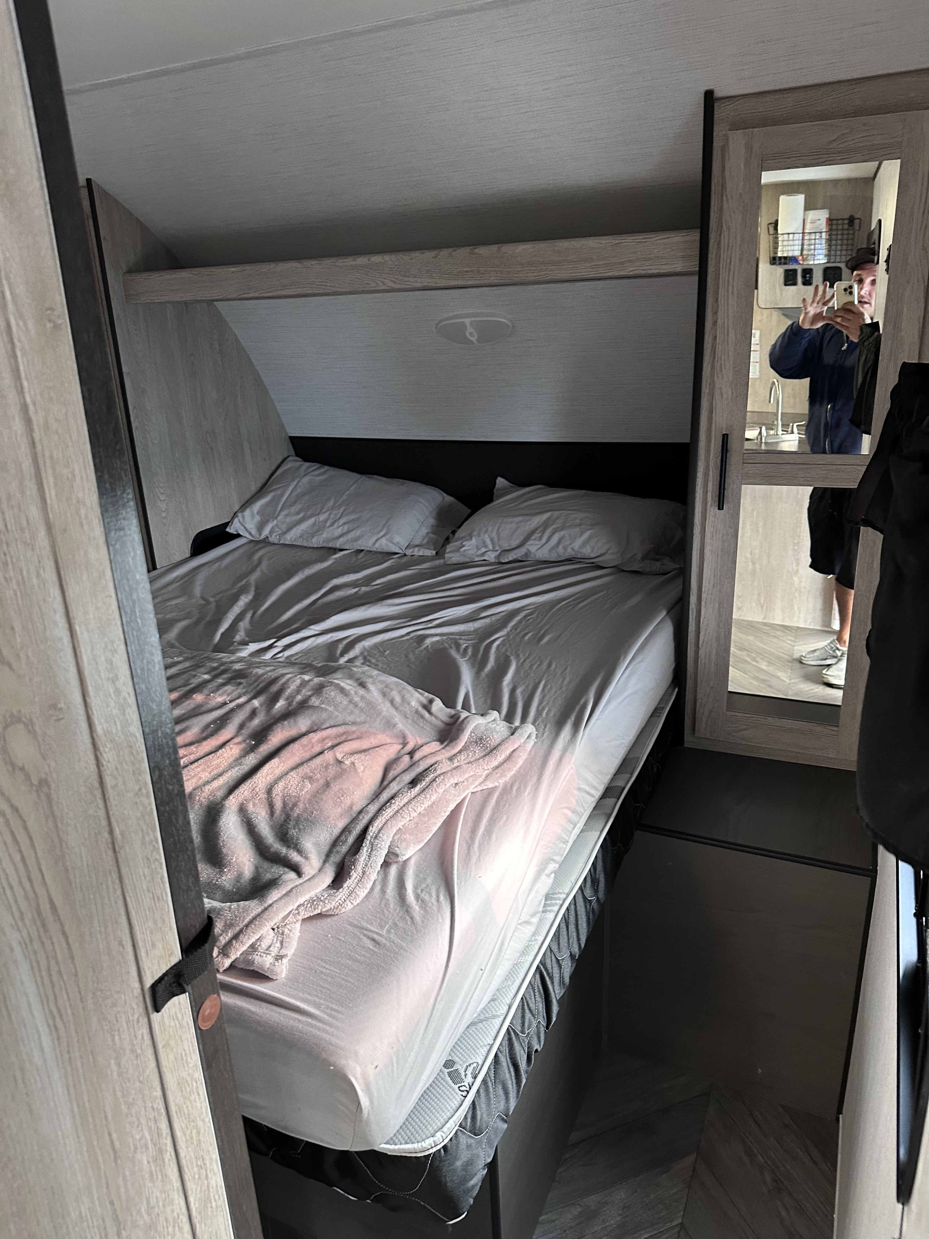 RV rental bedroom with a bed, light gray linens, and a mirror reflecting a person and a sink.. Heartland RVs Prowler 2022
