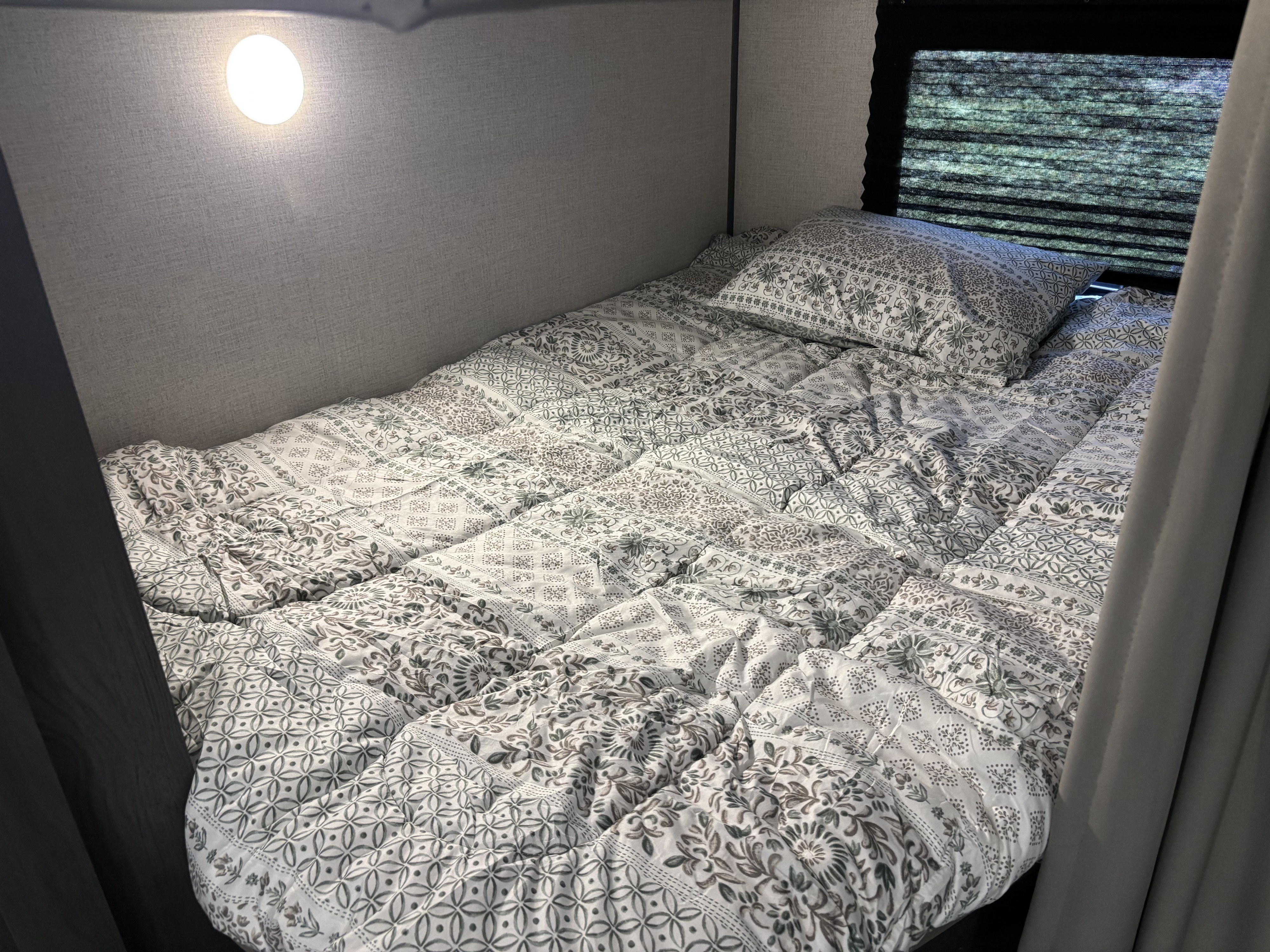 Comfortable RV rental bedroom featuring patterned bedding, an overhead lamp, and window with blinds.. Dutchmen Colorado Gold Rush Edition 2021