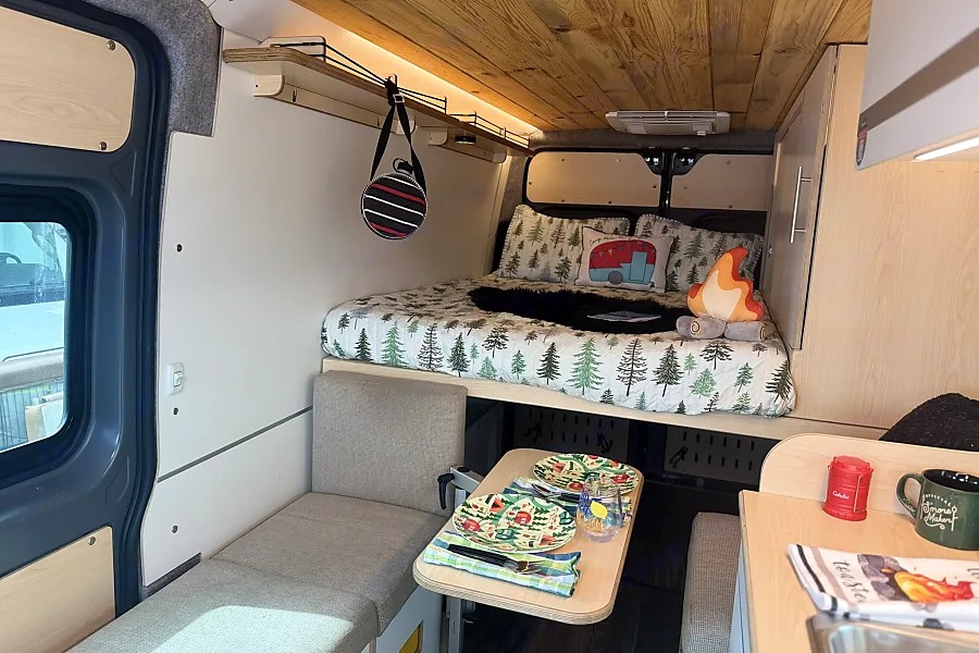 Cozy RV rental interior featuring a bed with nature-themed bedding, a dining table set for two, and wooden ceiling.. Dave & Matt Vans Dave & Matt Vans Class B 2023