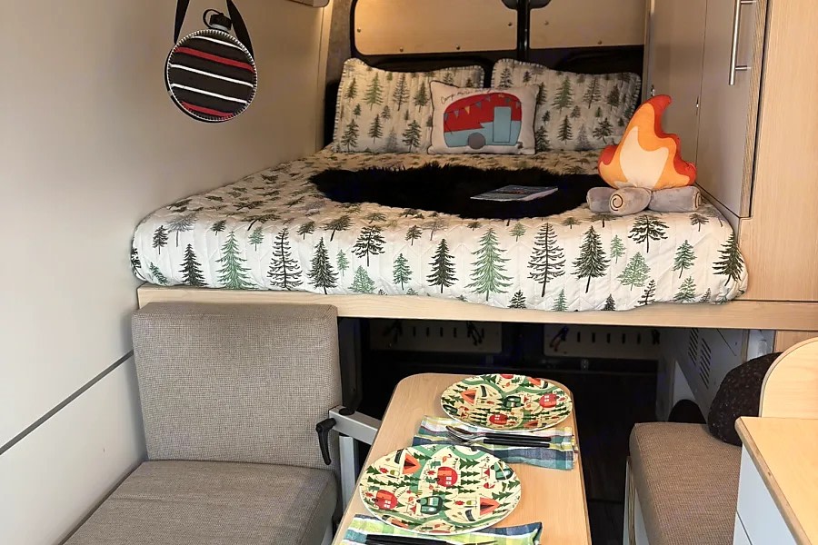 Inside a cozy rv rental: forest-patterned bed, camper pillow, campfire toy, & dining area with camping plates.. Dave & Matt Vans Dave & Matt Vans Class B 2023