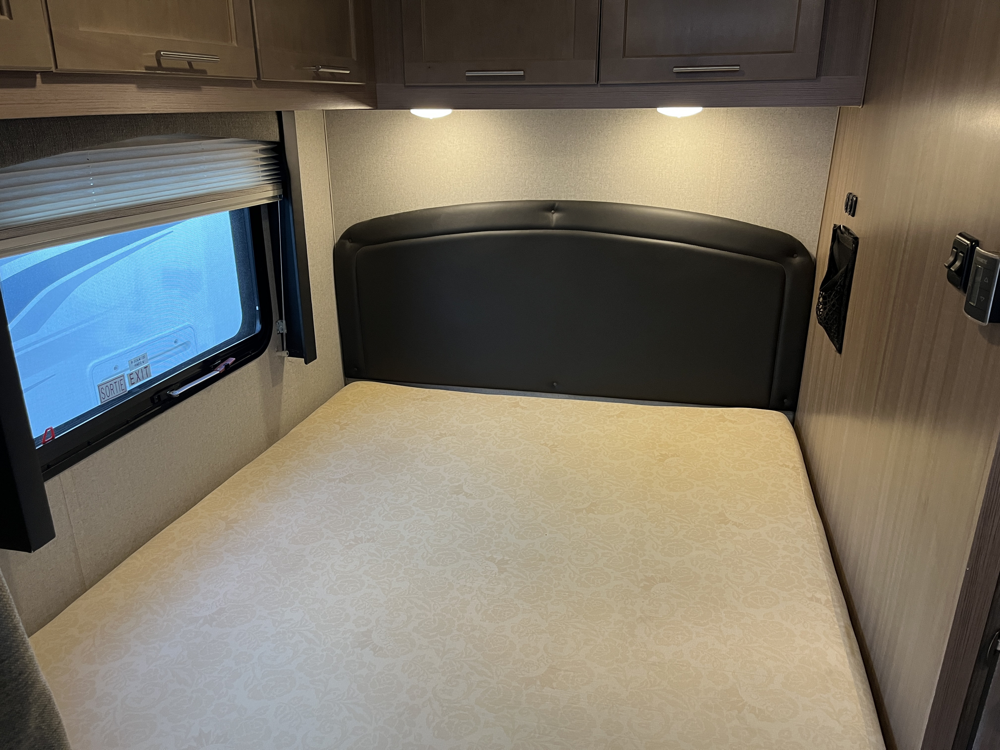 Cozy RV rental bedroom with patterned mattress, dark headboard, window, overhead cabinets, and wall controls. Ideal for travel.. Adventurer 23 RB 2021