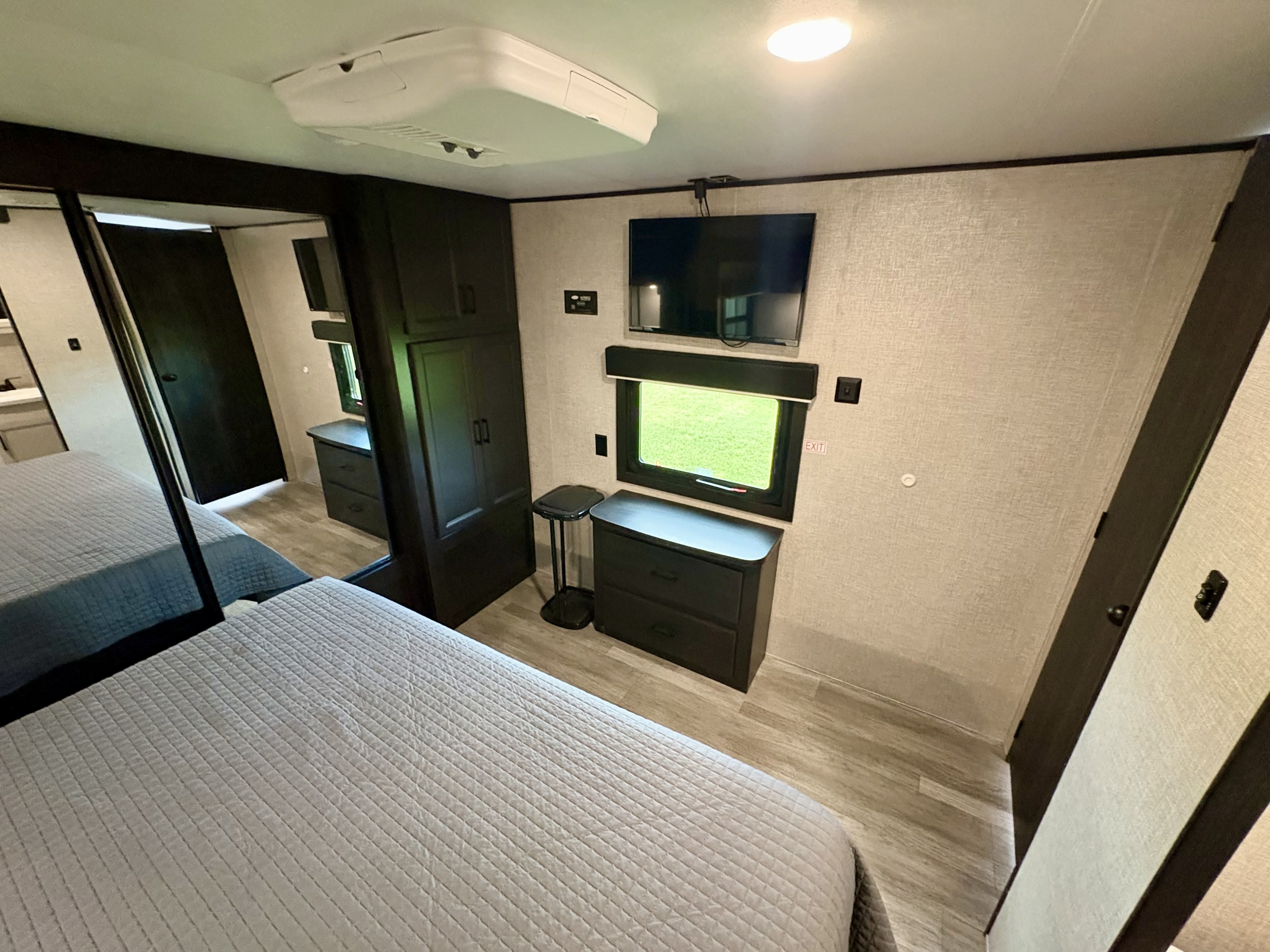 RV rental bedroom featuring a grey bed, dark wood wardrobe & dresser, wall-mounted TV, and window with a green view.. Jayco Jay Flight 2022