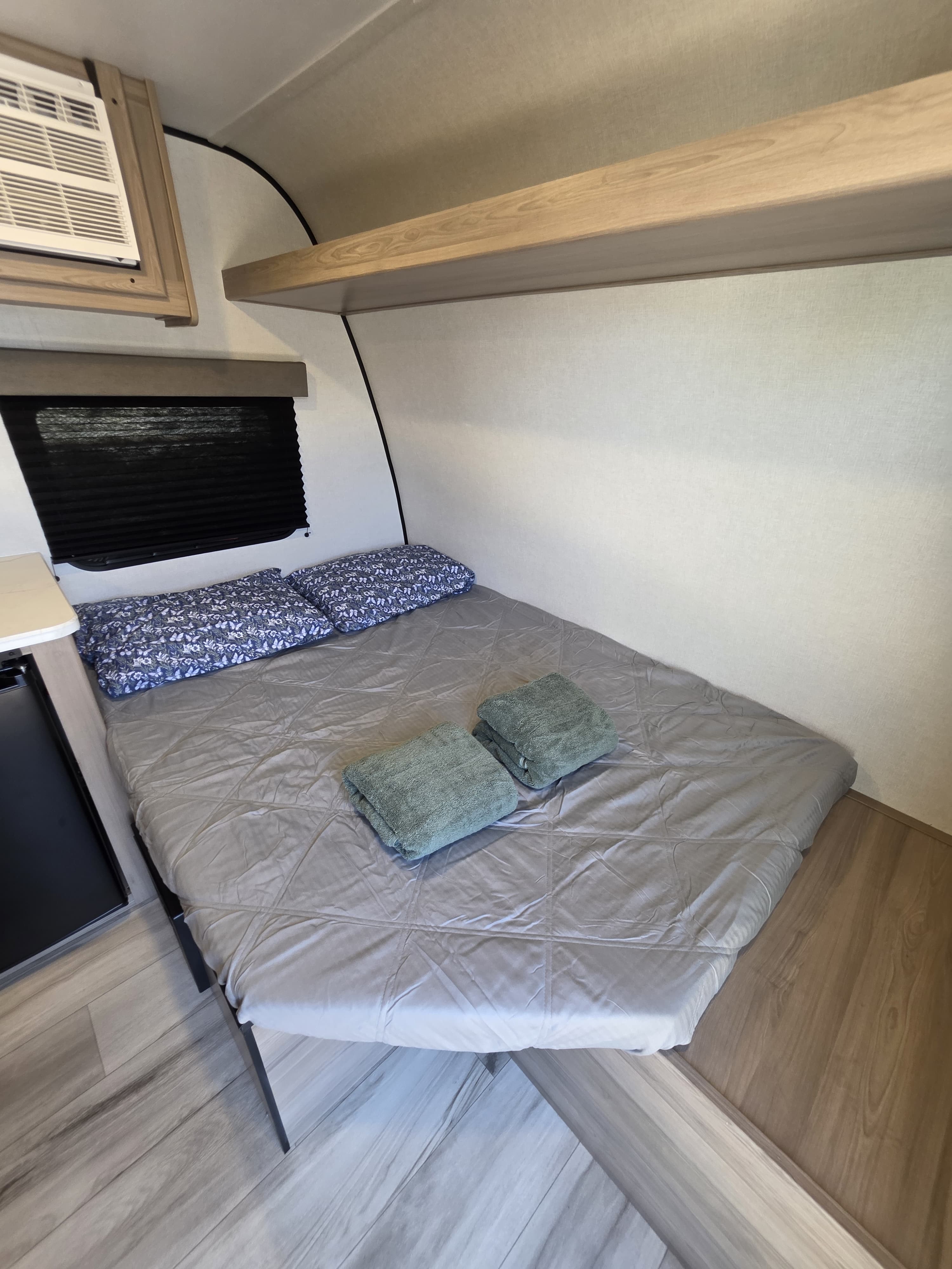Cozy RV rental bedroom with a compact bed, blue patterned pillows, and folded green towels. Features a window with blind and AC unit.. Keystone RV Coleman 13B 2026