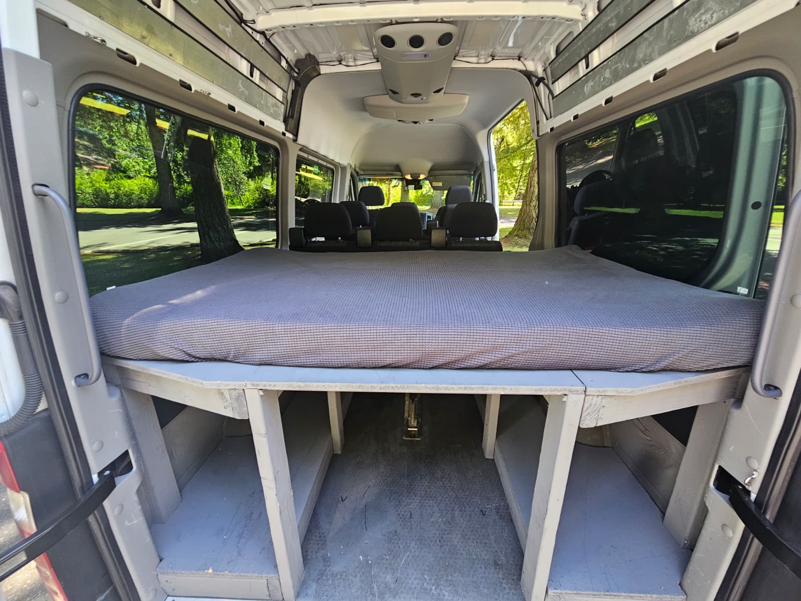 Interior of an RV rental camper van showing a raised bed with storage underneath, looking towards the front seats.. Mercedes-Benz Sprinter 2016