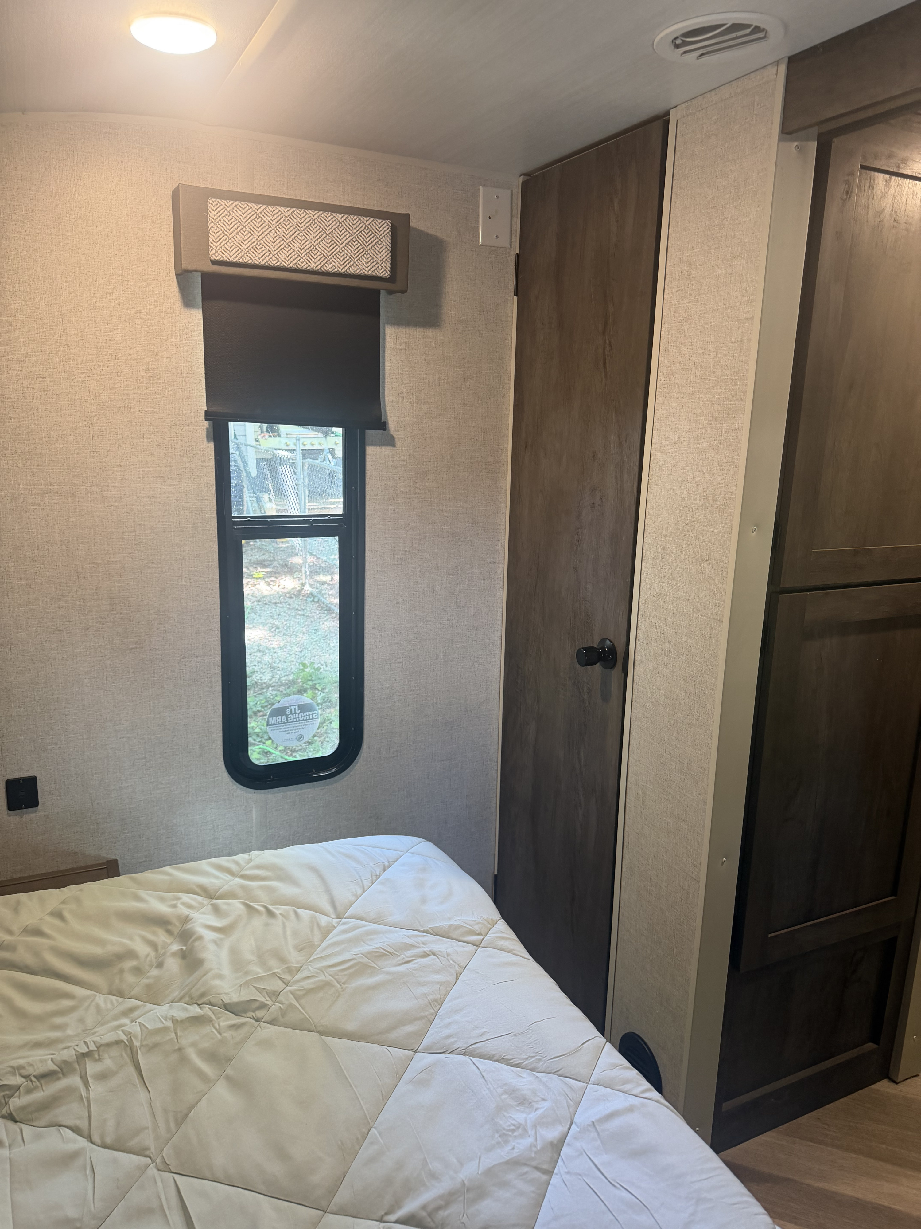 This rv rental bedroom features a textured wall, window with blind, white comforter, dark wood cabinetry, ceiling light, and AC vent.. Forest River Wildwood X-Lite 2021
