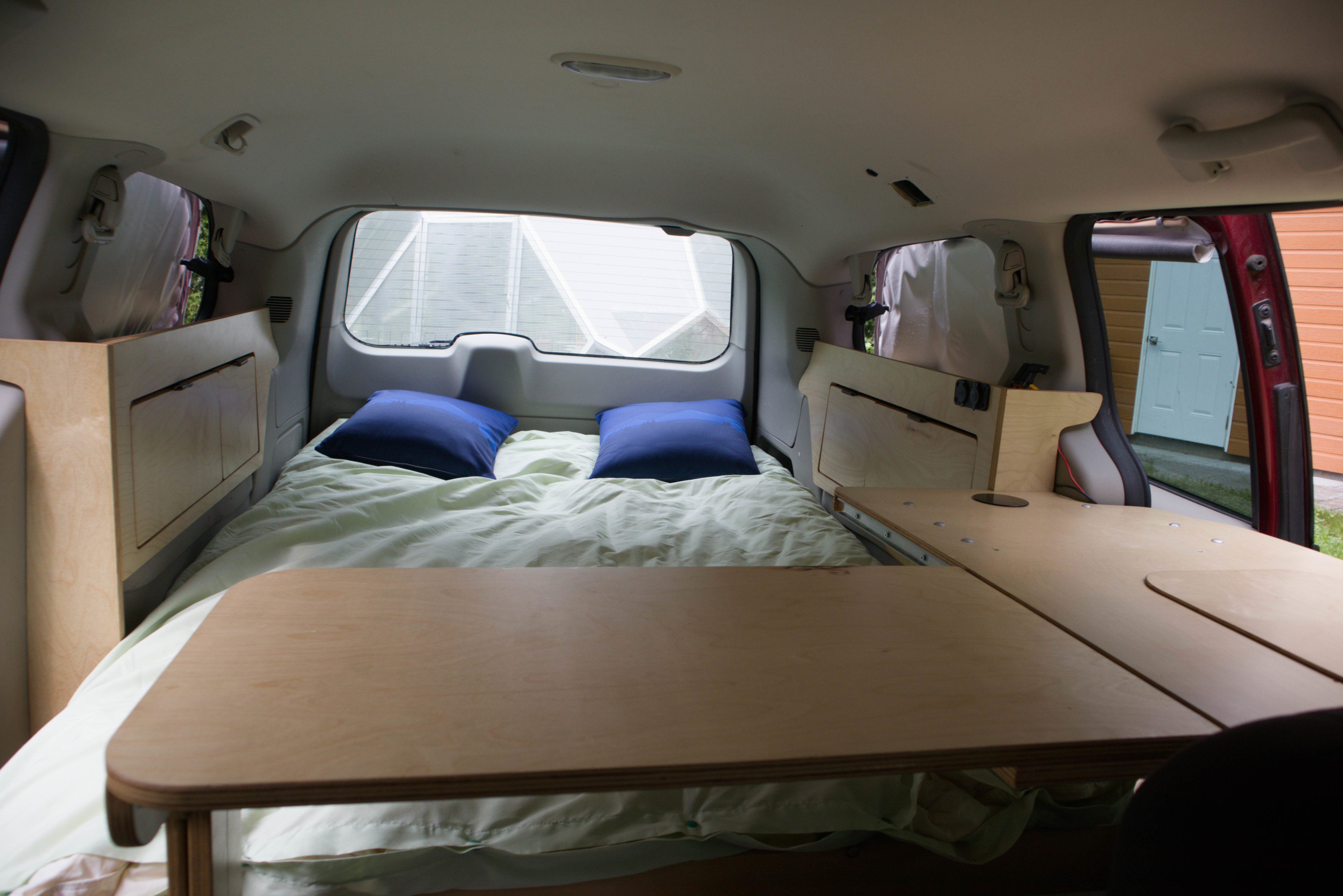 RV rental campervan interior with a cozy bed, blue pillows, and custom wooden buildout including storage and a table for travel.. Dodge Grand Caravan 2015