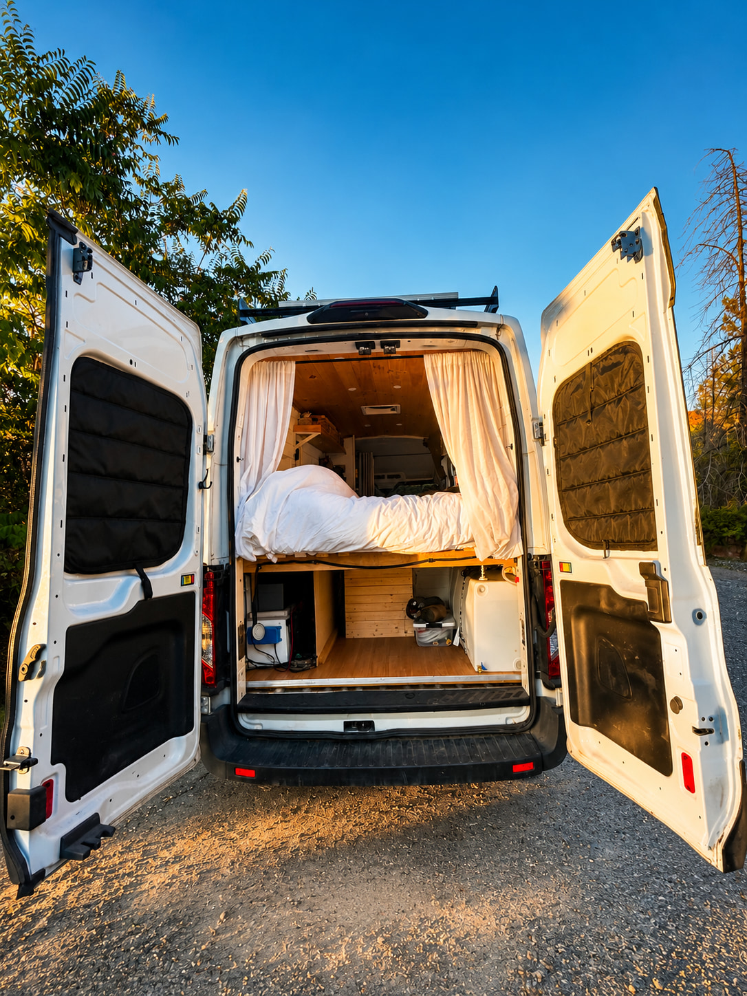RV rental: Rear view of a white camper van with open doors, revealing a cozy bed, wooden interior, and organized storage.. Ford Transit 2020