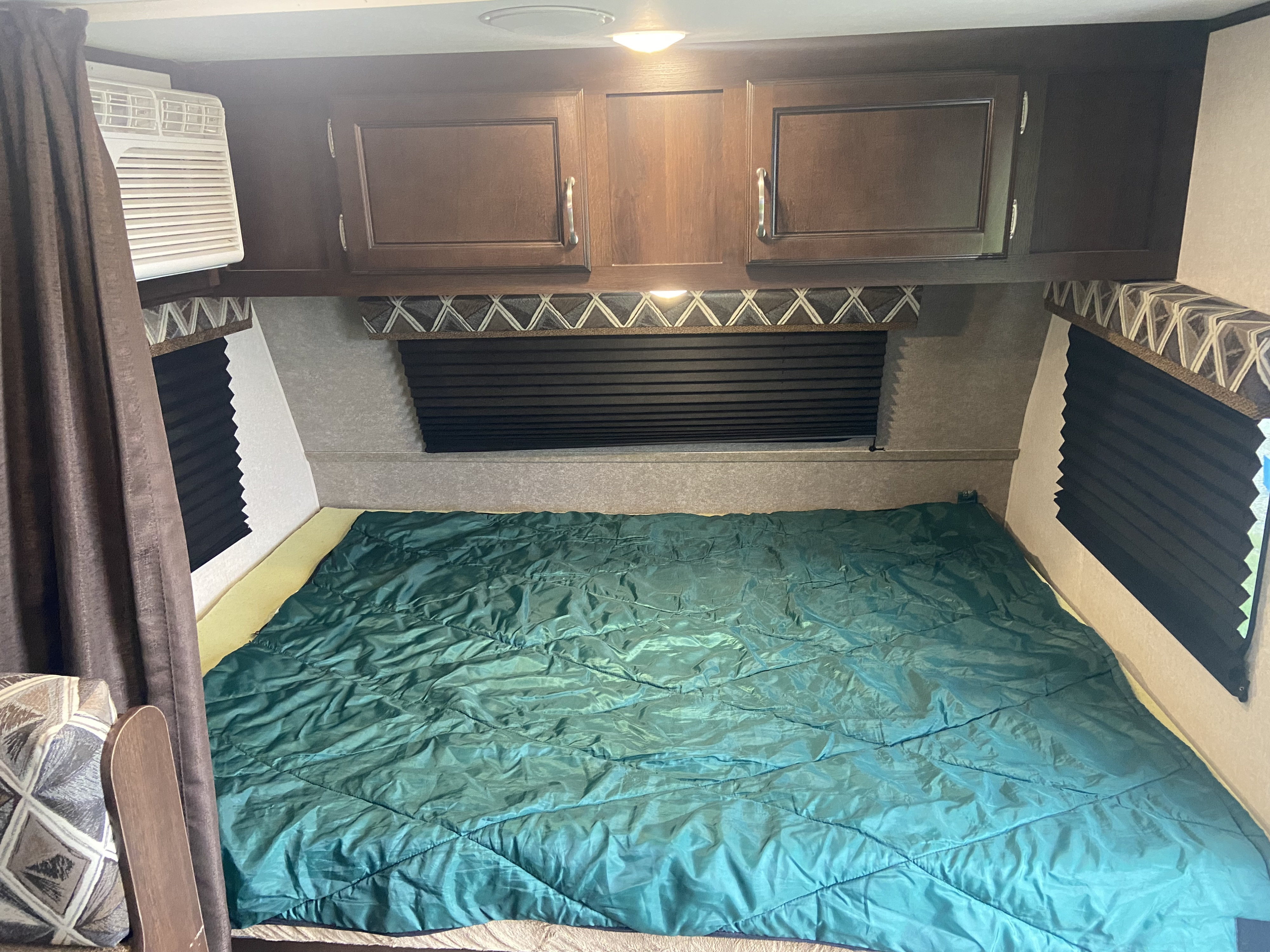 Cozy bed area in an RV rental, featuring a green comforter, overhead cabinets, AC unit, and window blinds.. Jayco Jay Flight SLX 2016