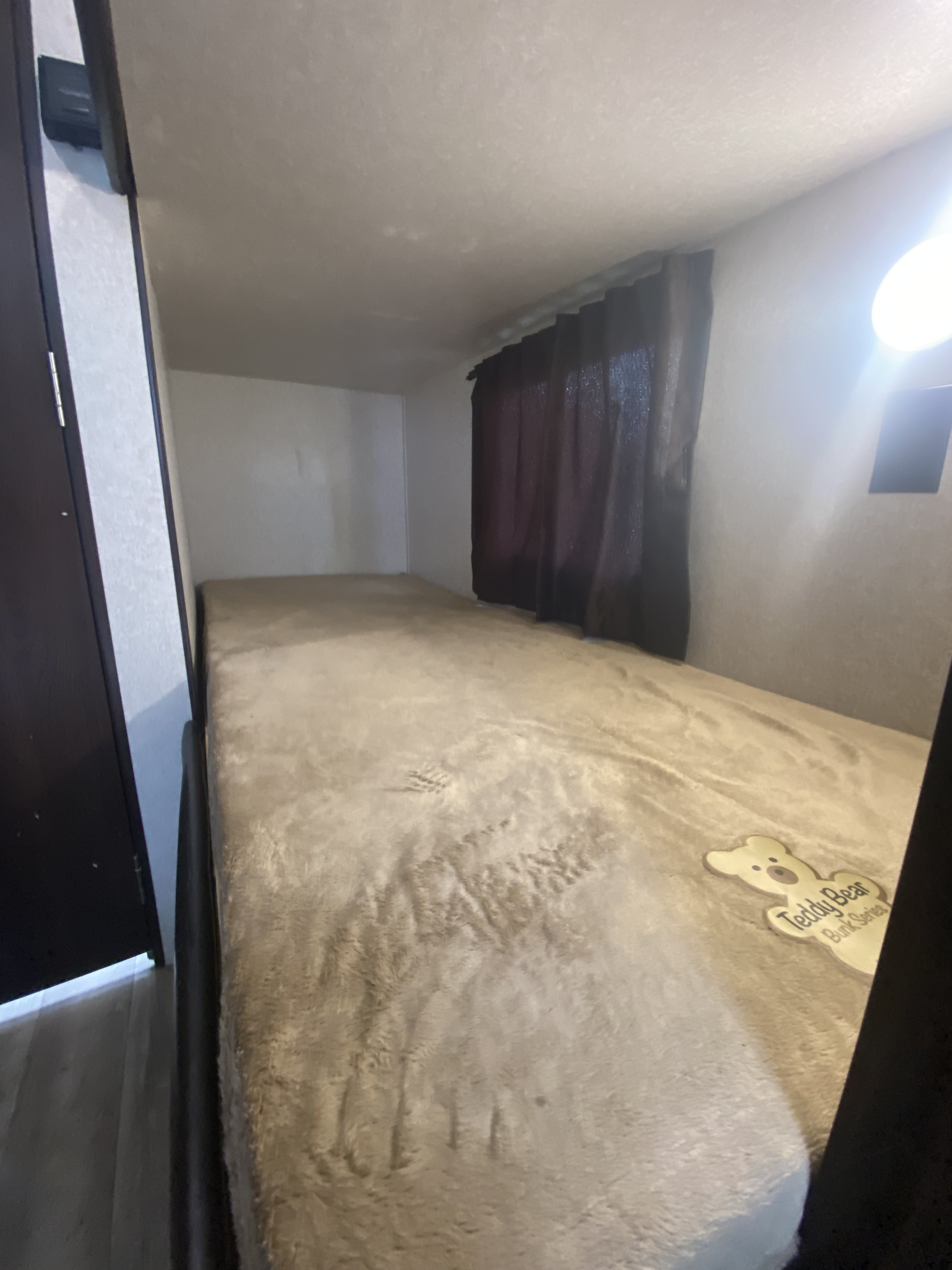 This RV rental features a cozy bunk bed with a soft, light-colored blanket and a "Teddy Bear Bunk Series" tag. Dark curtains cover a window, and a bright light illuminates the wall.. Jayco Jay Flight SLX 2016
