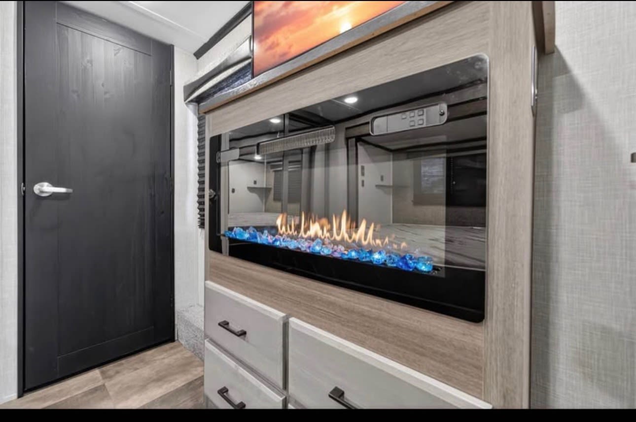 Modern RV rental interior featuring a stylish electric fireplace with blue glass, a TV, and storage drawers, beside a dark door.. Crossroads RV Cameo 2022