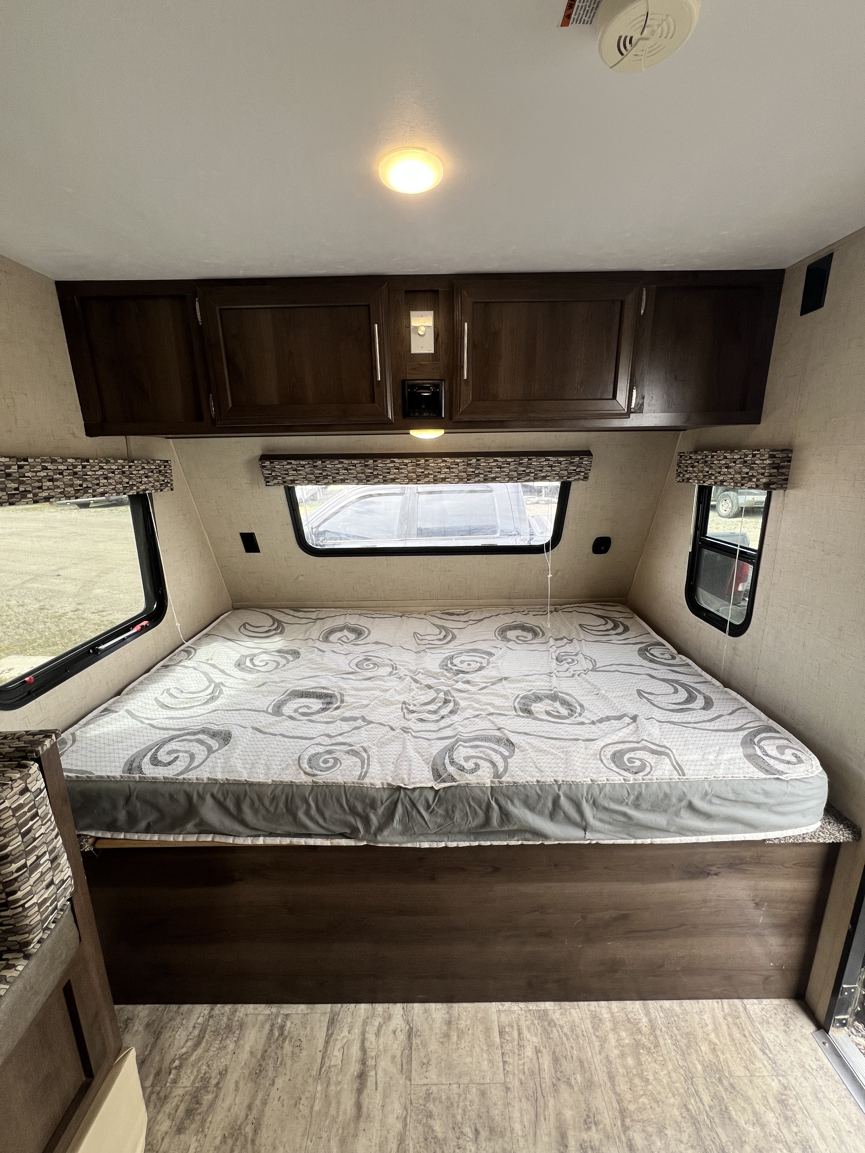 Inside an RV rental: a neat bedroom with a spacious bed, overhead cabinets, windows, and ceiling lighting.. KZ Sportsmen Classic 181BH 2018