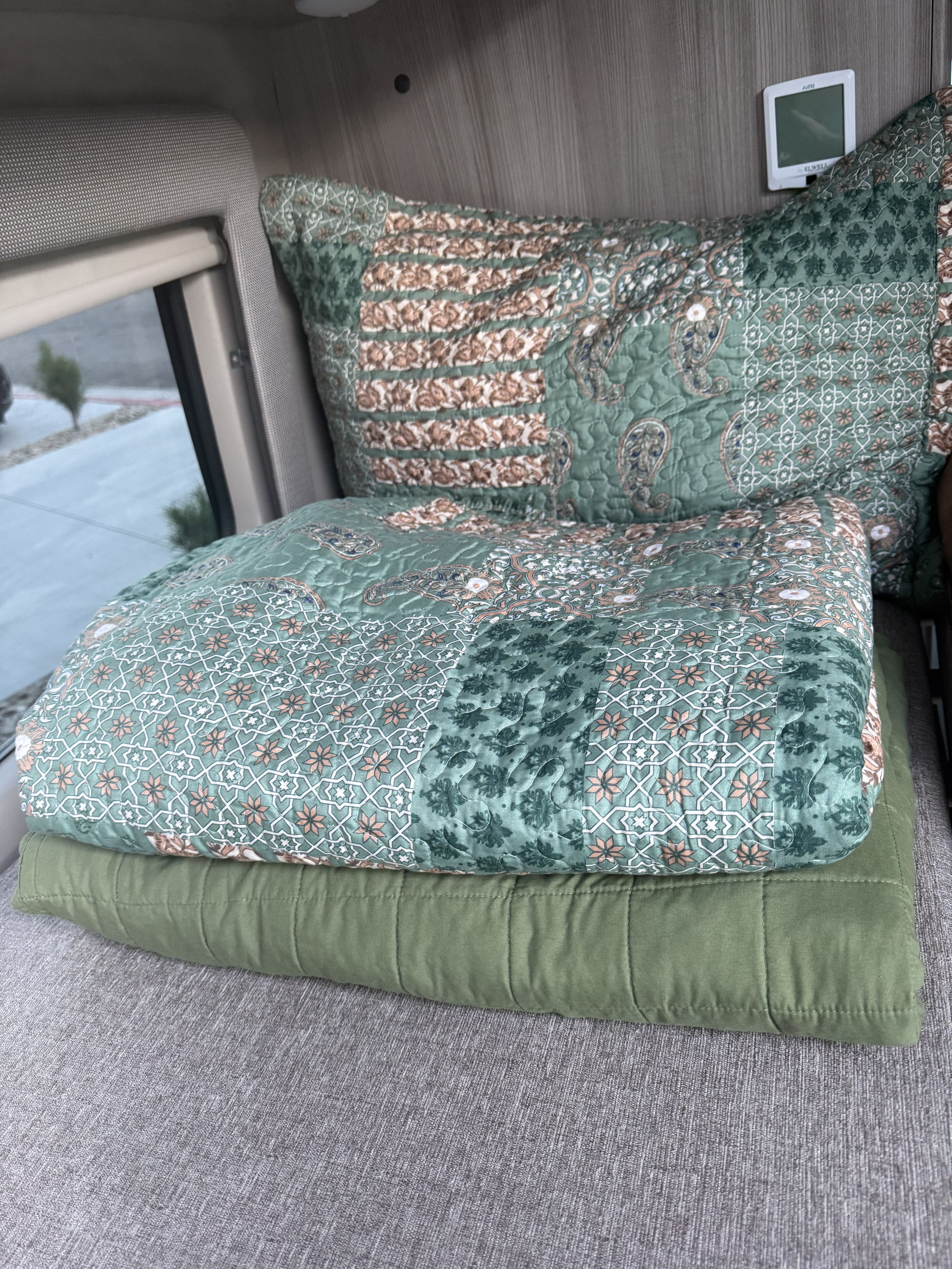 In an RV rental, patterned green and brown quilted bedding and pillows are neatly stacked, beside a window and small thermostat.. Thor Motor Coach 2023 Thor Motor Coach Scope 18A 2023