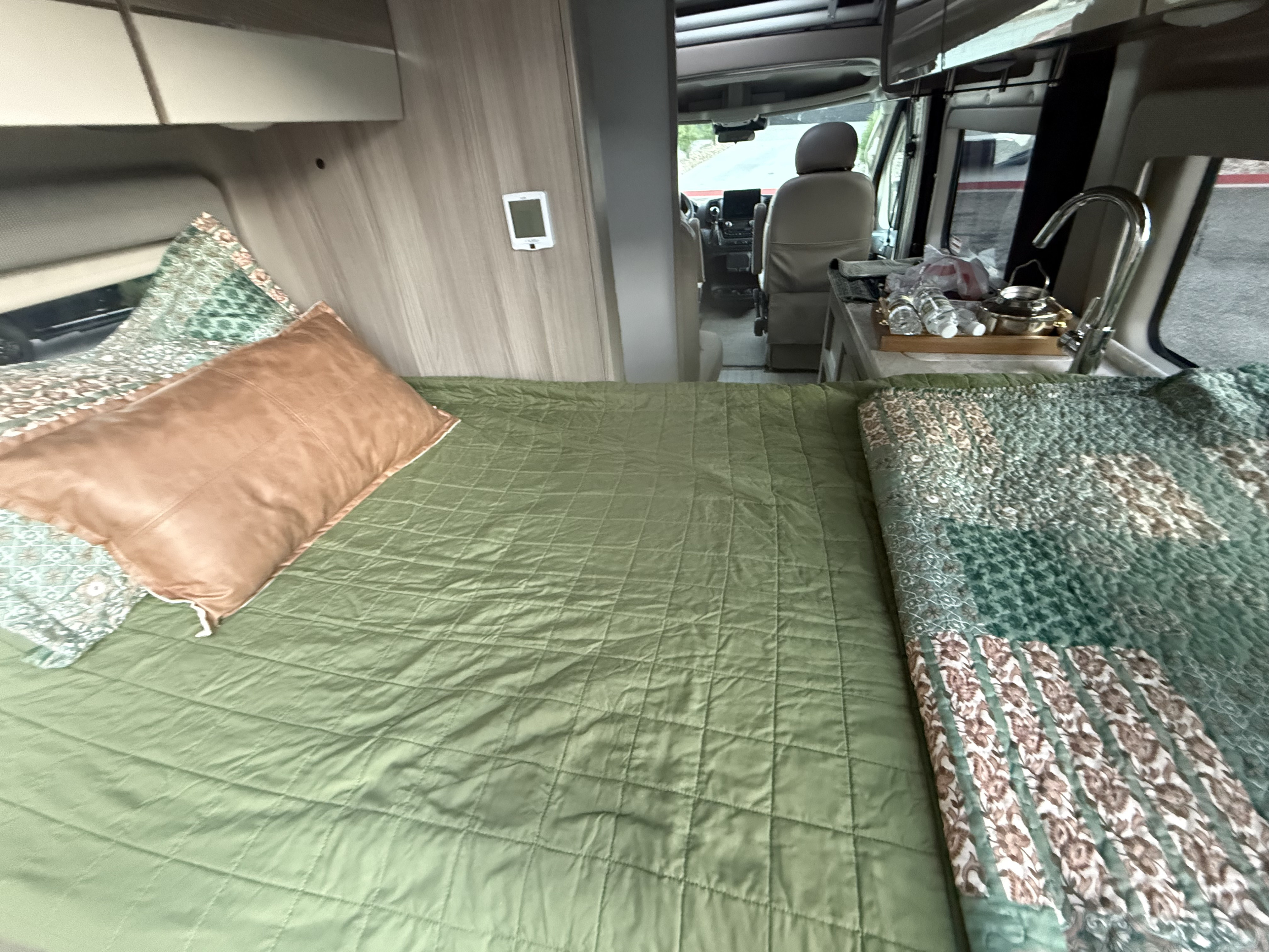 Inside an RV rental, showing a bed with green bedding, brown pillow, and a small kitchen area near the driver's seat.. Thor Motor Coach 2023 Thor Motor Coach Scope 18A 2023