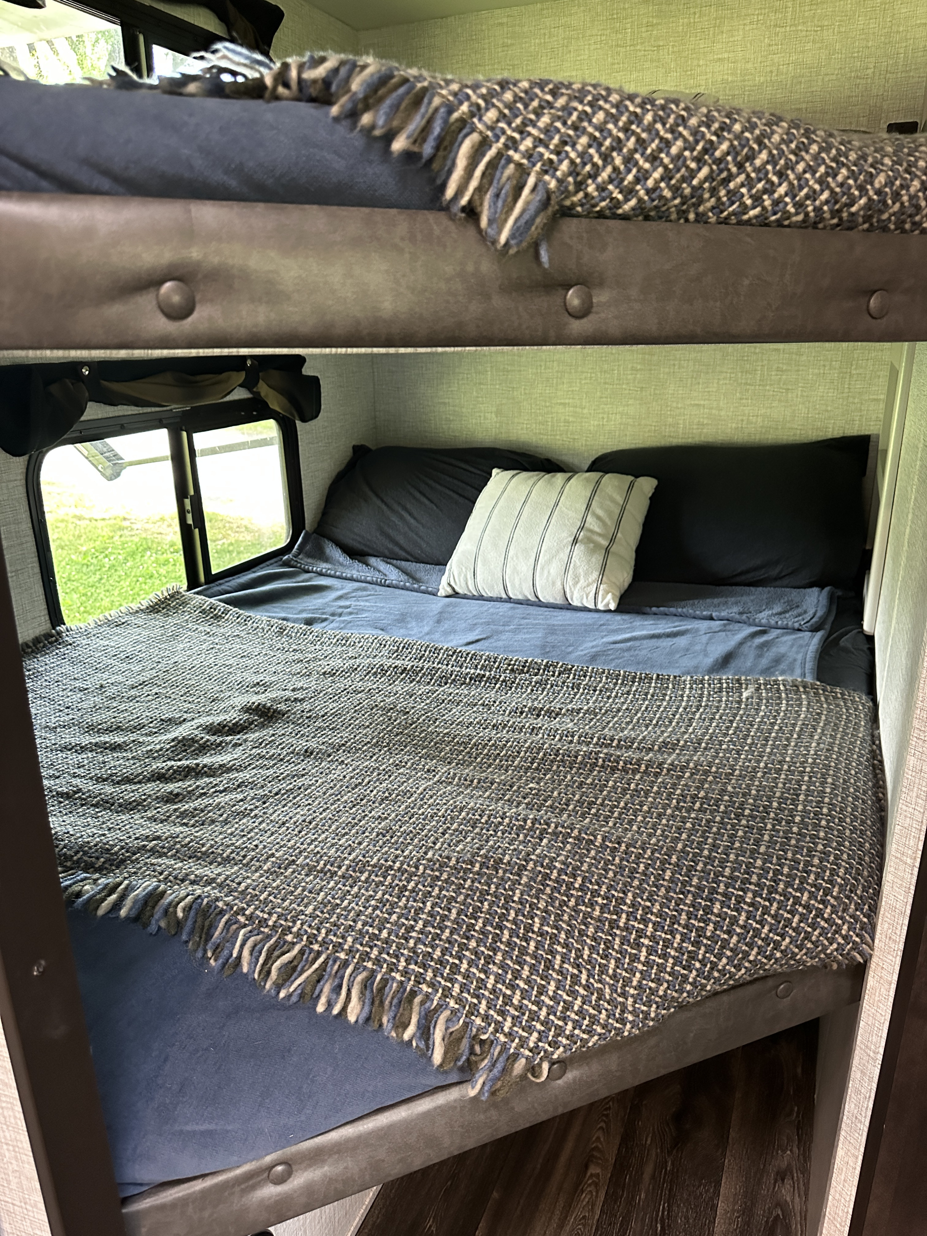 Cozy RV rental bunk beds: blue bedding, patterned throws. Lower bunk has pillows and a window view of green grass.. Keystone RV Bullet Crossfire 2021
