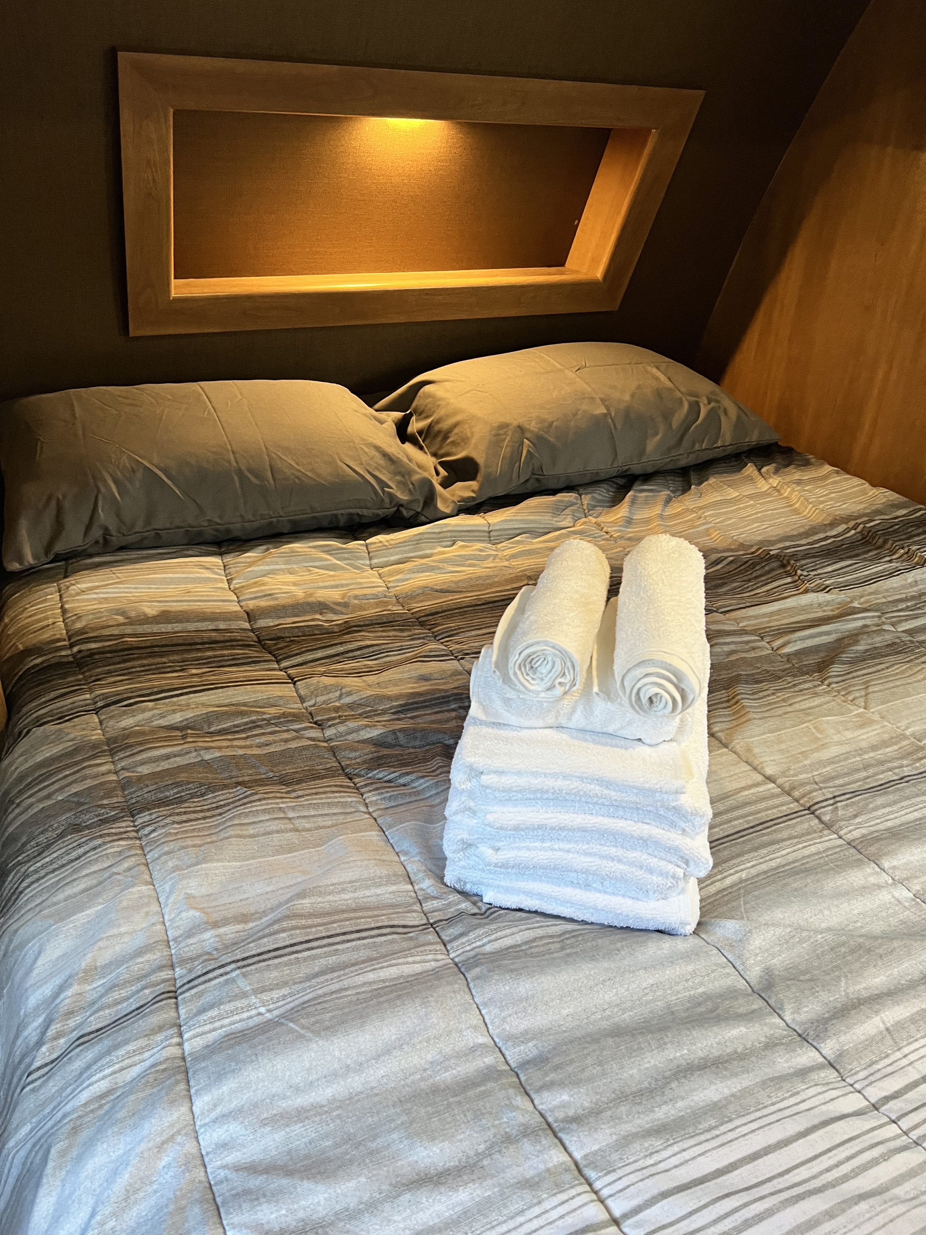 RV rental's cozy sleeping area features striped bedding, gray pillows, and a stack of fresh white towels for comfort.. Coachmen Apex 2018