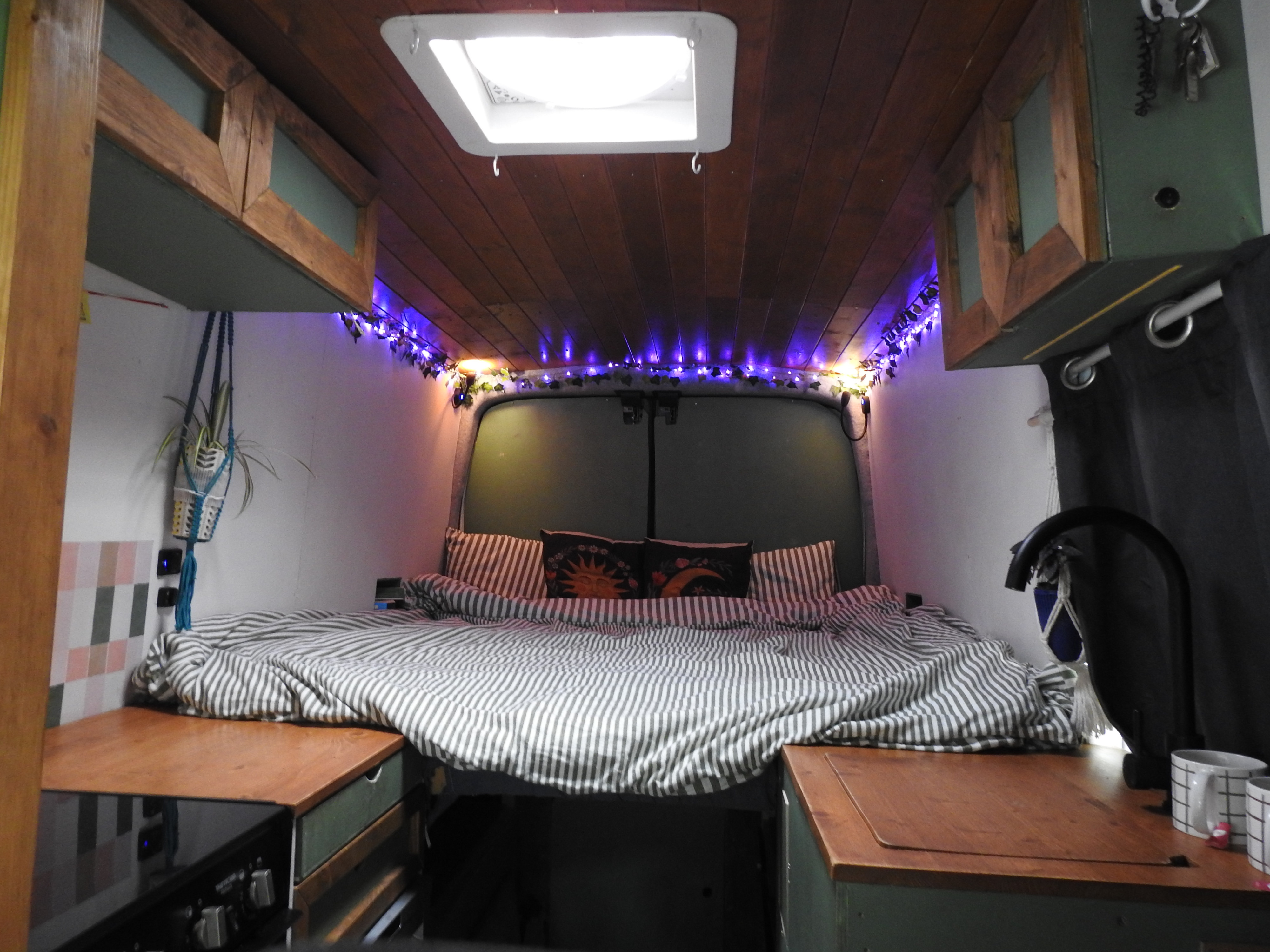 Cozy camper van rental interior. Features a bed with striped bedding, wood ceiling, fairy lights, and compact kitchen/storage.. Ford Transit 2018