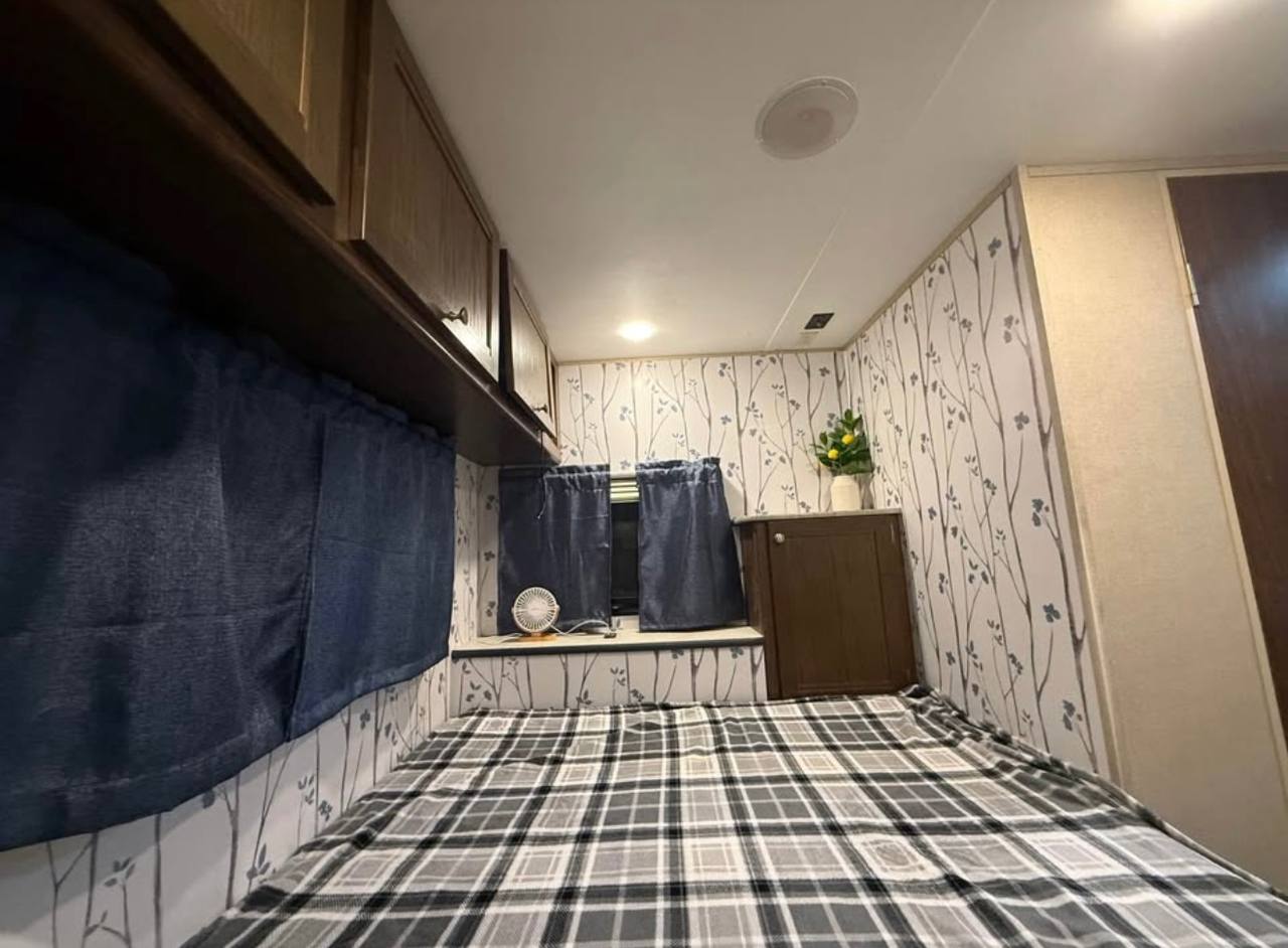 Cozy RV rental bedroom with plaid bedding, patterned walls, blue curtains, and overhead cabinets for a comfortable stay.. Keystone RV coleman lantern 17RD 2017